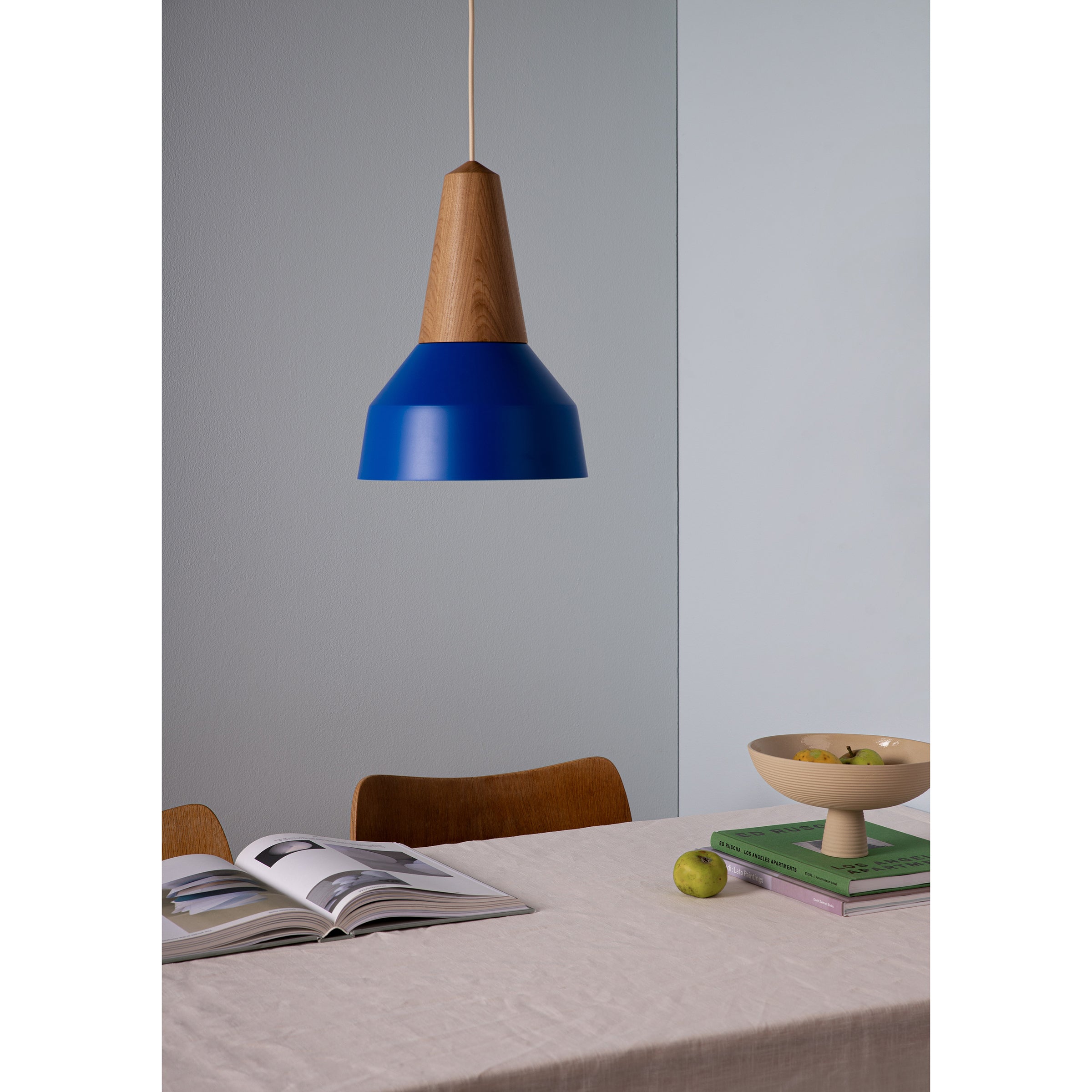 A modern dining area features the Schneid Eikon Basic True Blue pendant light, crafted from certified wood, above a table. The table holds an open book, a green apple, stacked books, and a fruit-filled wooden bowl. Chairs are set against the light-colored wall in the background.