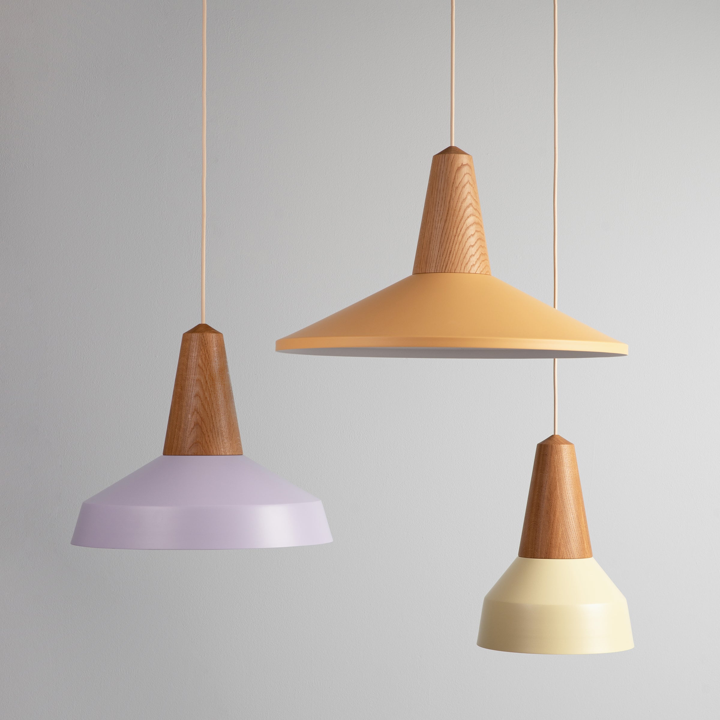 Three Schneid Eikon Basic Wax pendant lights with certified wood tops and matte shades in pastel purple, orange, and yellow hang at varying heights against a neutral gray background.