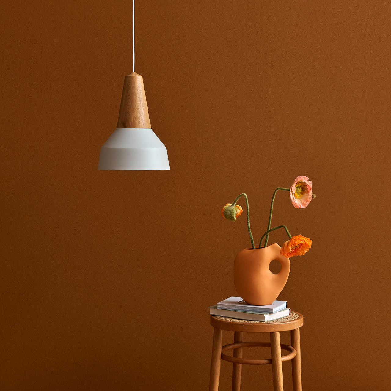 A Schneid Eikon Basic White modern pendant light hangs over a wooden stool made from certified wood. The stool supports books and a unique terracotta vase with three colorful flowers, set against a warm brown wall.