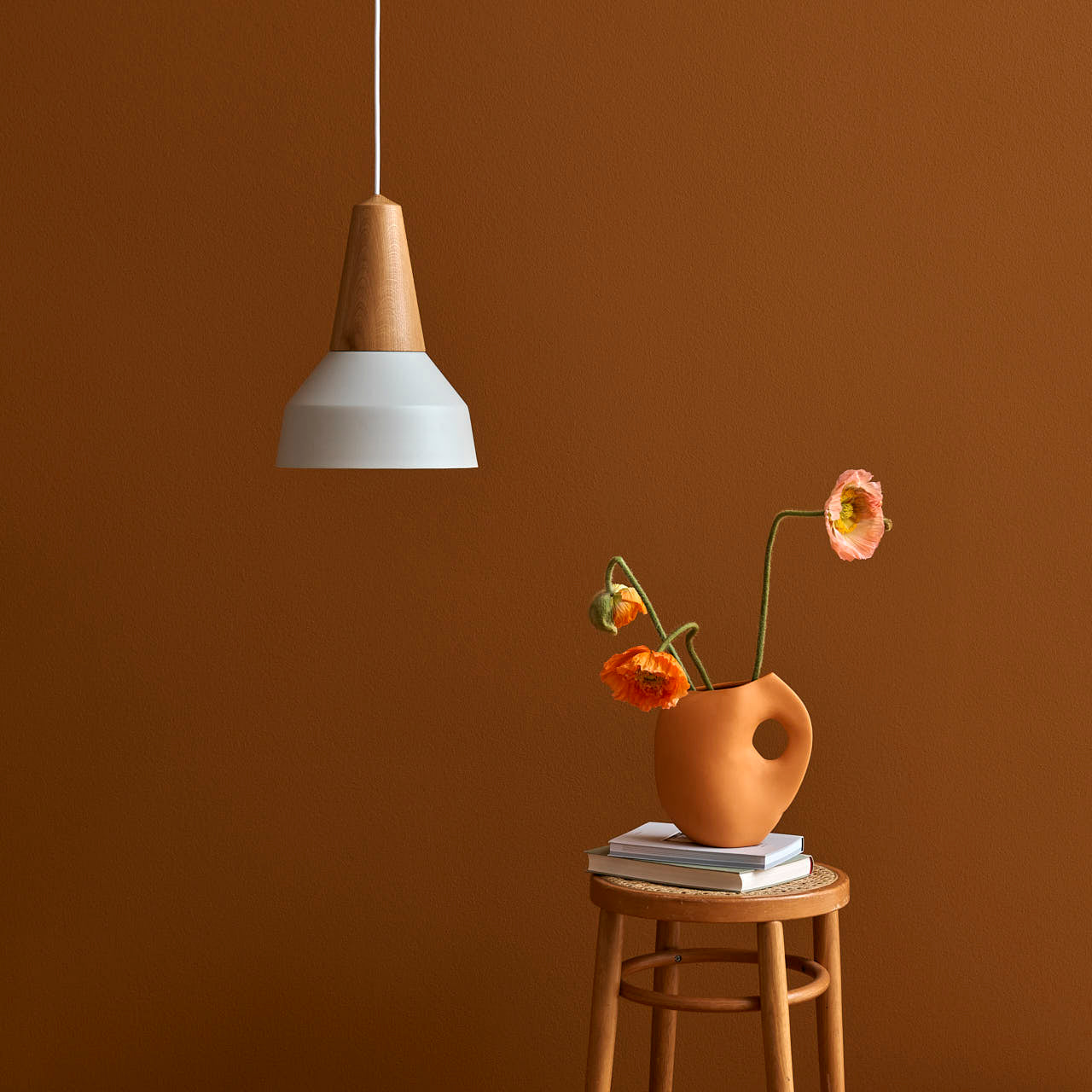 The Schneid Eikon Basic True Blue pendant lamp with a modern design hangs above an orange vase filled with poppies on a small wooden stool made from certified wood, all set against a warm brown wall that enhances the cozy atmosphere.