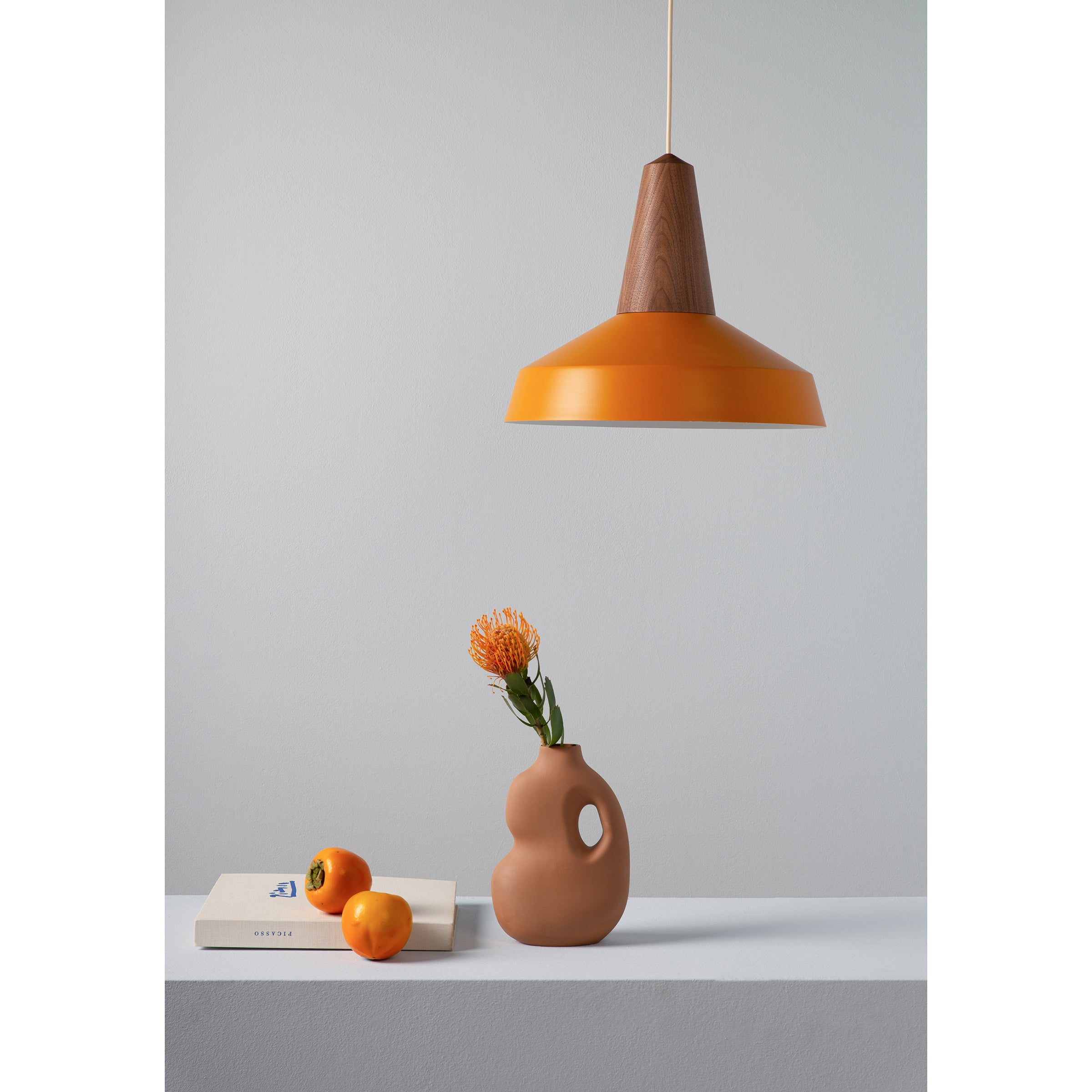 The minimalist scene highlights Schneids Eikon Circus Turmeric pendant lamp above a white surface. Beneath, a sculptural vase with an orange flower is flanked by two small oranges and a closed beige book, set against a plain gray wall, epitomizing contemporary expression.