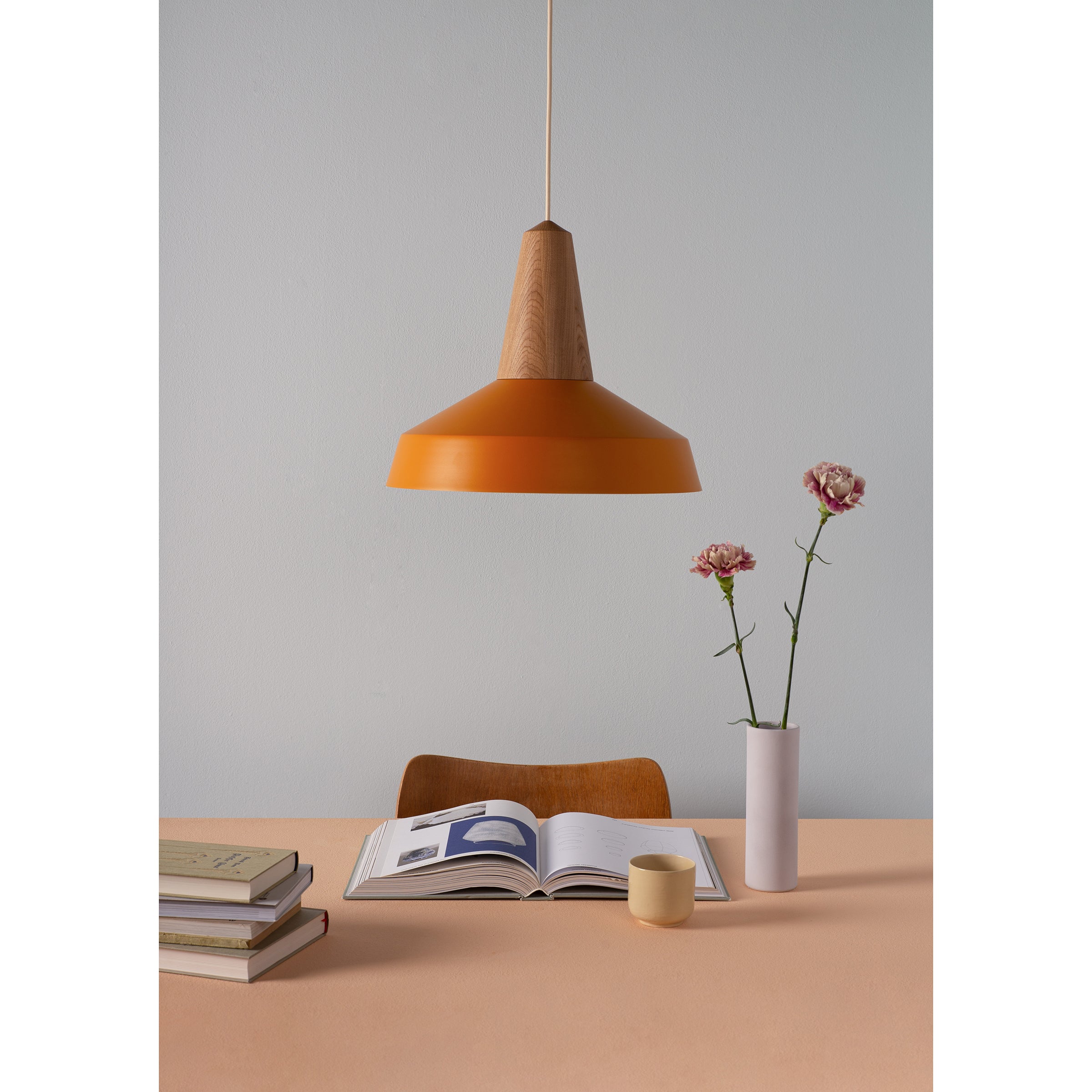 A minimalist scene features a Schneid Eikon Circus Turmeric pendant lamp hanging above a table adorned with a stack of books, an open magazine, a small beige cup, and a vase with two pink flowers. A partially visible wooden chair adds to the spaces contemporary feel.