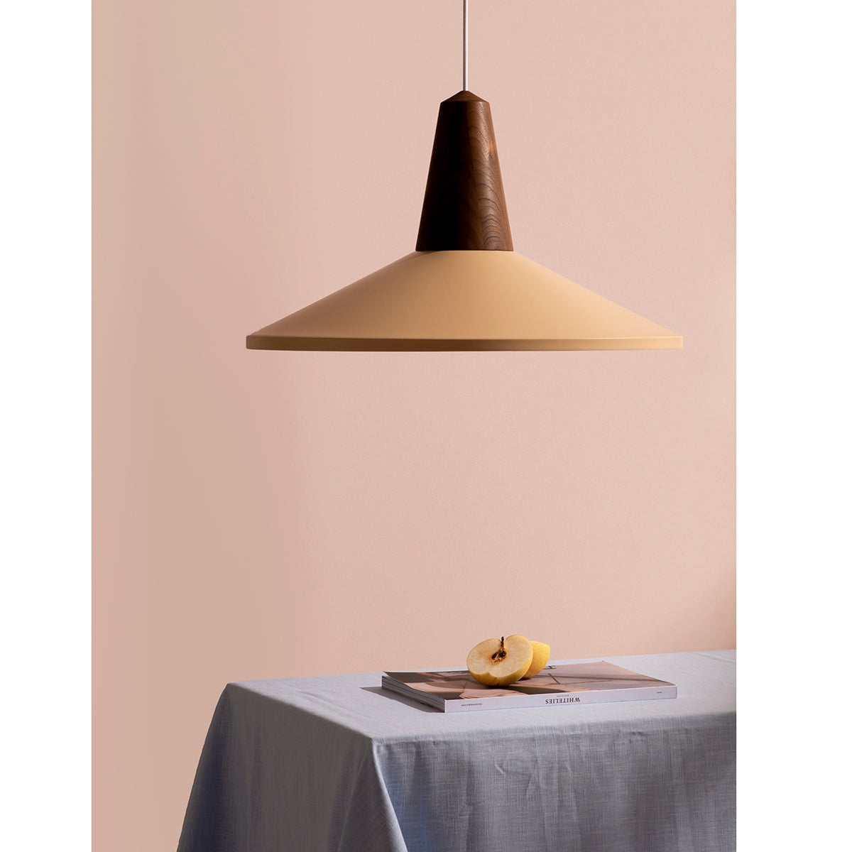 Schneids Eikon Shell Peach Sherbet lamp elegantly hangs above a table draped in light gray cloth. A magazine and a sliced apple with exposed seeds add charm against the beige backdrop, highlighting this sophisticated pendant lighting.