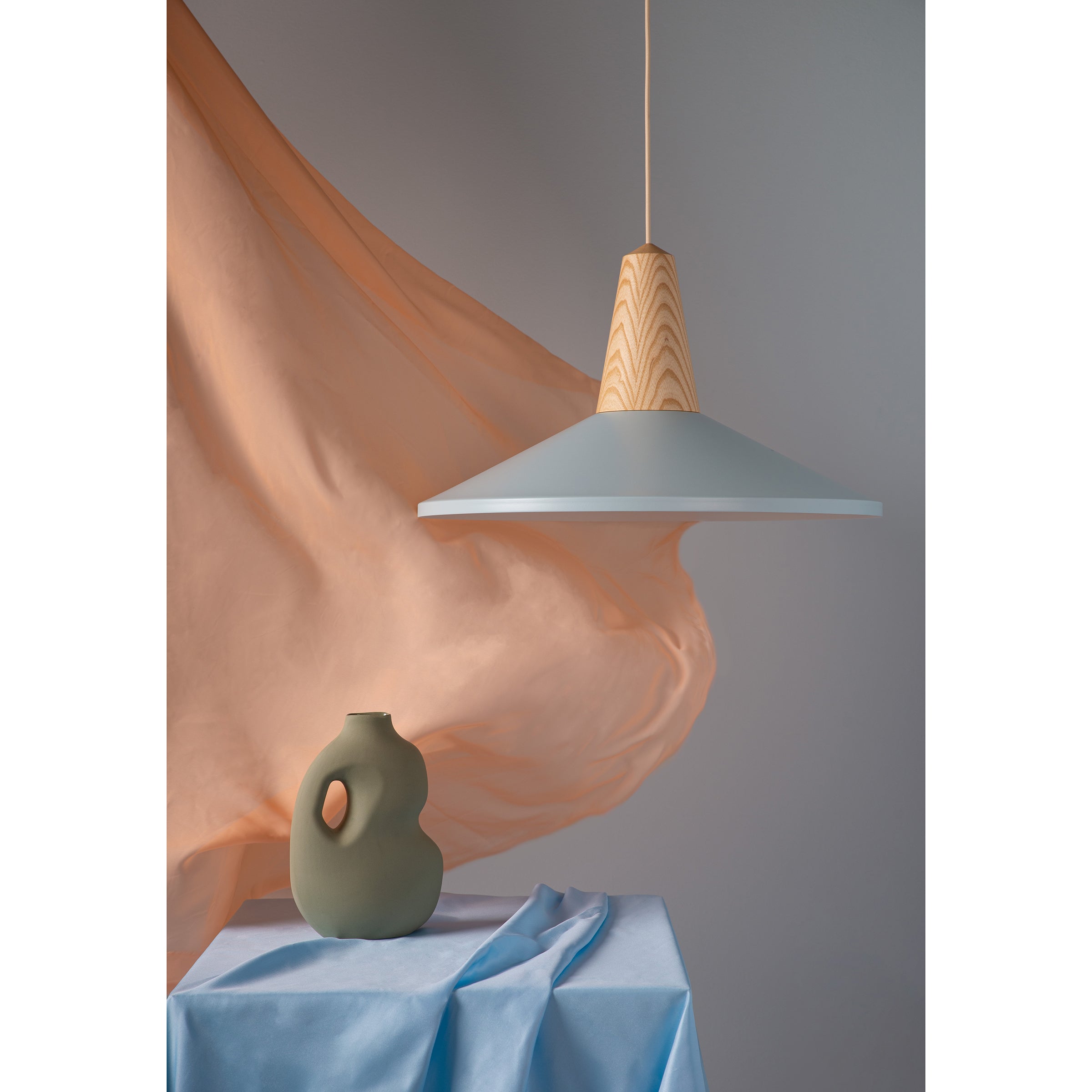 The scene features Eikon Shell Sky Blue pendant lighting by Schneid with a pastel lamp above. Below, a uniquely shaped vase is on a table draped in pale blue fabric, while peach-hued fabric billows in the background for a stylish, elegant look.
