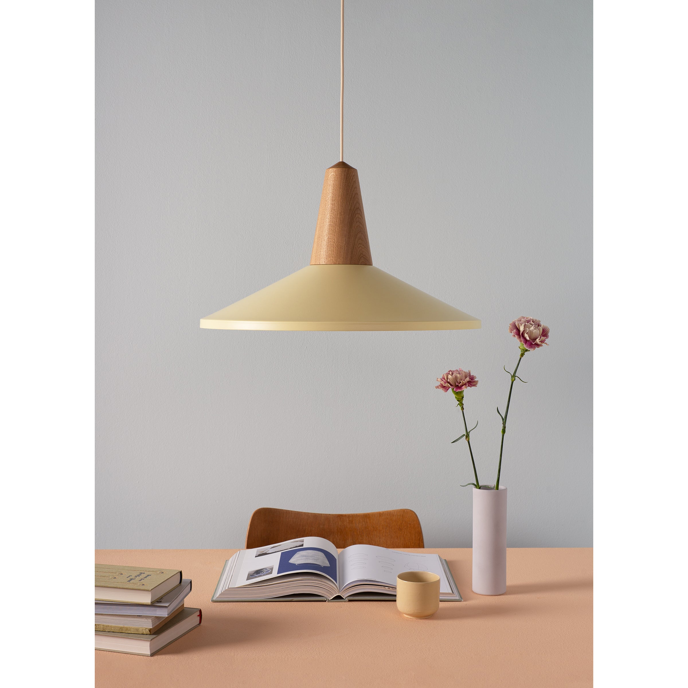 A minimalist scene features the Eikon Shell Wax beige pendant lamp from Schneid, casting light on an open book. Stacked books, a small cup, and a vase with two pink flowers rest nearby. A simple chair crafted from certified wood is positioned at the table.