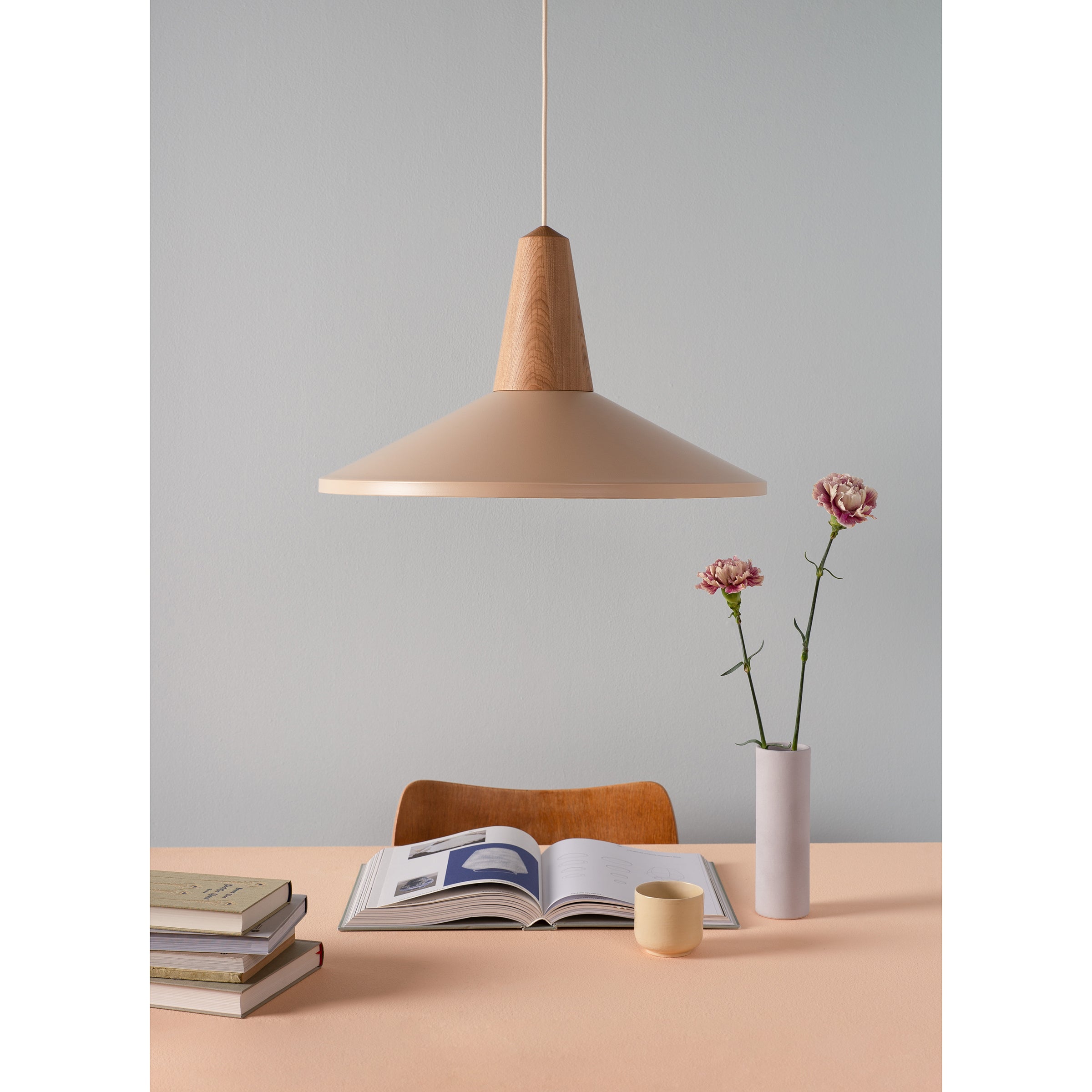 A minimalist scene features a wooden table with an open book, stacked books, and a vase with two pink carnations. Above, the Schneid Eikon Shell Wheat lampshade hangs, casting light on the setup with its magnetic system against a soft gray wall backdrop.