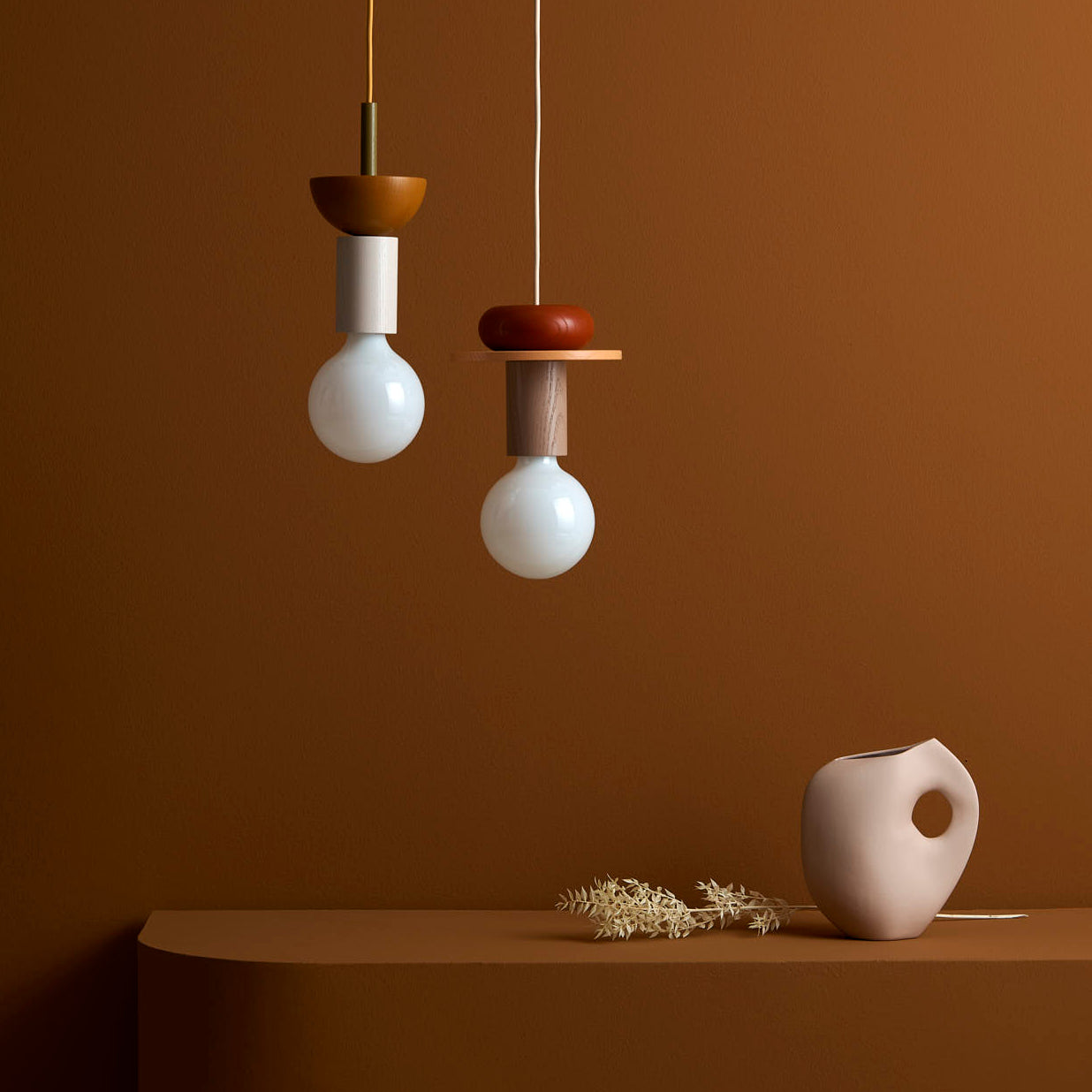 Two Junit Pan modular pendant lamps by Schneid hang over a curved, minimalist table against a warm brown backdrop. A small beige vase with dried flowers sits on the table, creating a cozy ambient light and stylish atmosphere.