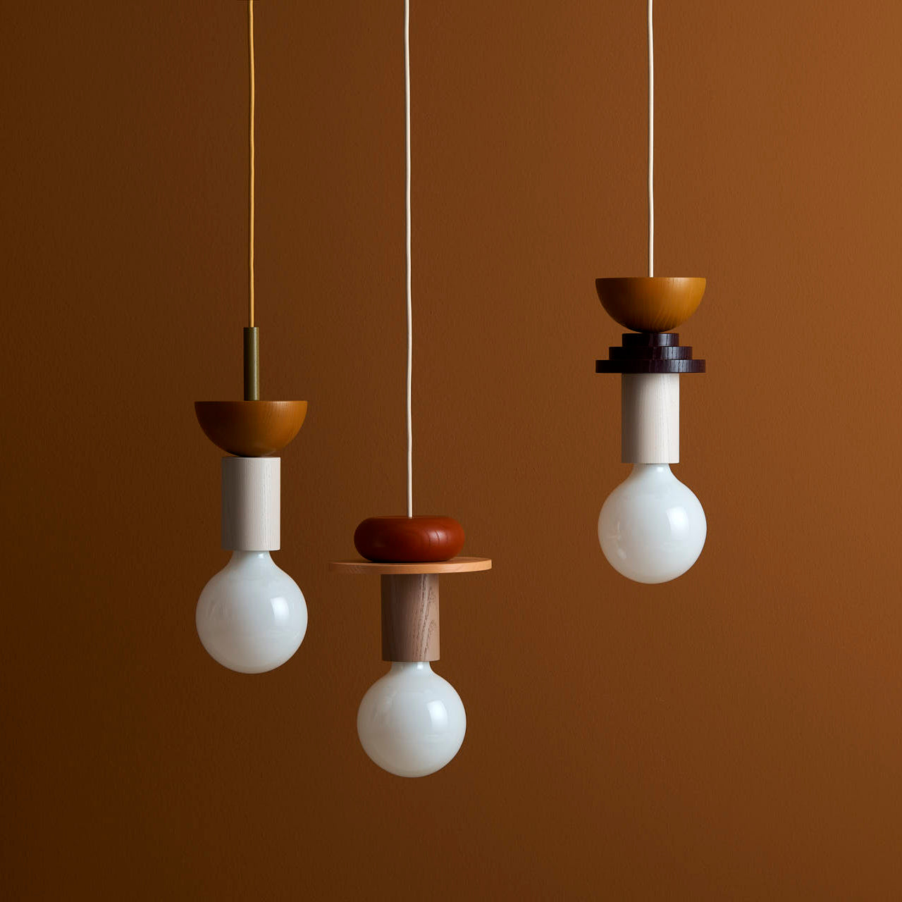 Set against a warm brown background, the Schneid Junit Pan pendant lights elegantly feature round white bulbs with geometric wooden fixtures made of ash wood and vibrant cords, offering ambient light in a minimalist and stylish design.