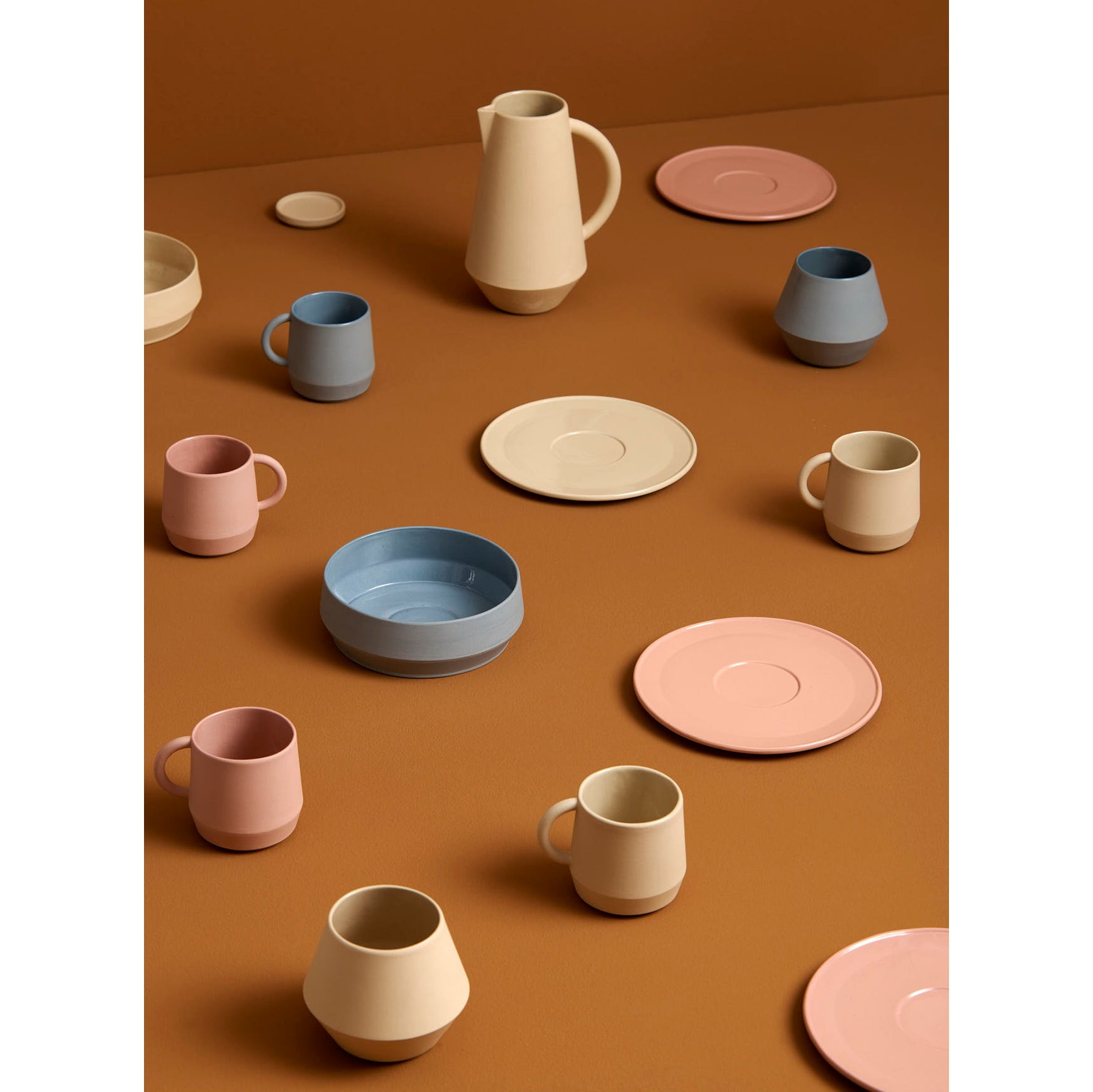 A stackable collection of Schneids Unison Small Plate and assorted ceramic tableware in soft pastels, featuring handmade ceramics such as mugs, plates, bowls, and a pitcher, arranged on a warm brown surface.