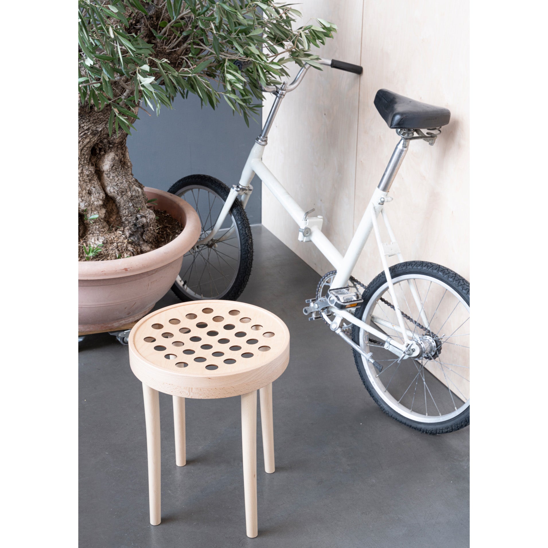 The Stool 822 by TON, featuring circular perforations, is beside a white bicycle with small wheels against a wooden wall. A large potted plant adds greenery, capturing Scandinavian minimalism and the refined touch of TONDesigners design.