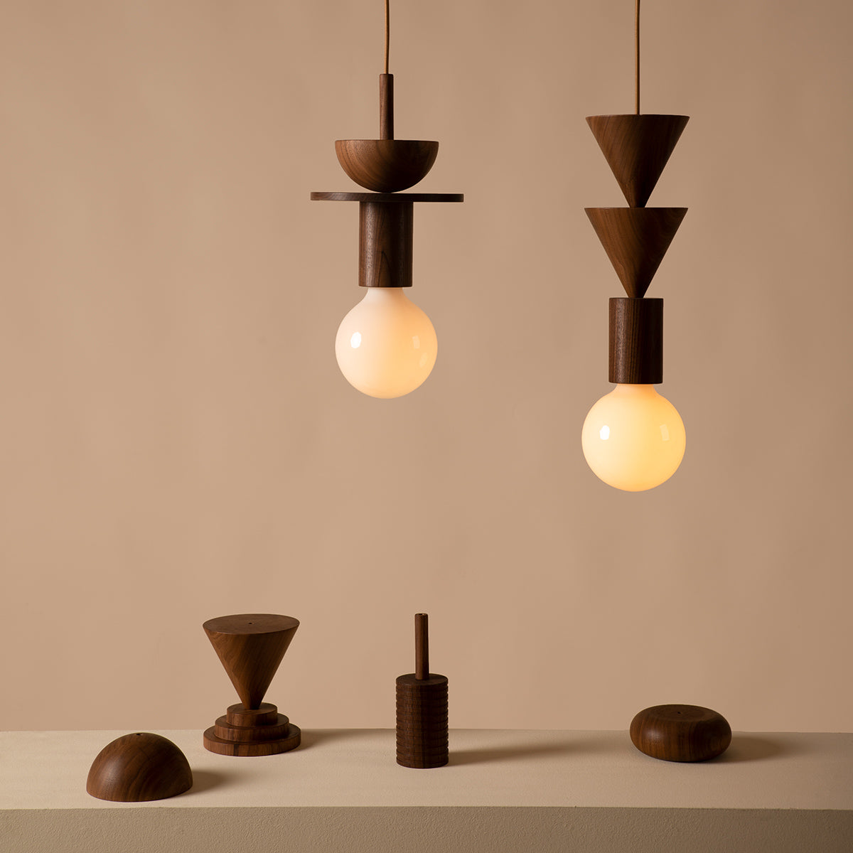 Two Junit Copla pendant lights by Schneid, featuring wooden geometric Bauhaus designs, hang over a table adorned with walnut sculptures: a cone, a handled cylinder, and an oval. The scene exudes a minimalist and earthy aesthetic.