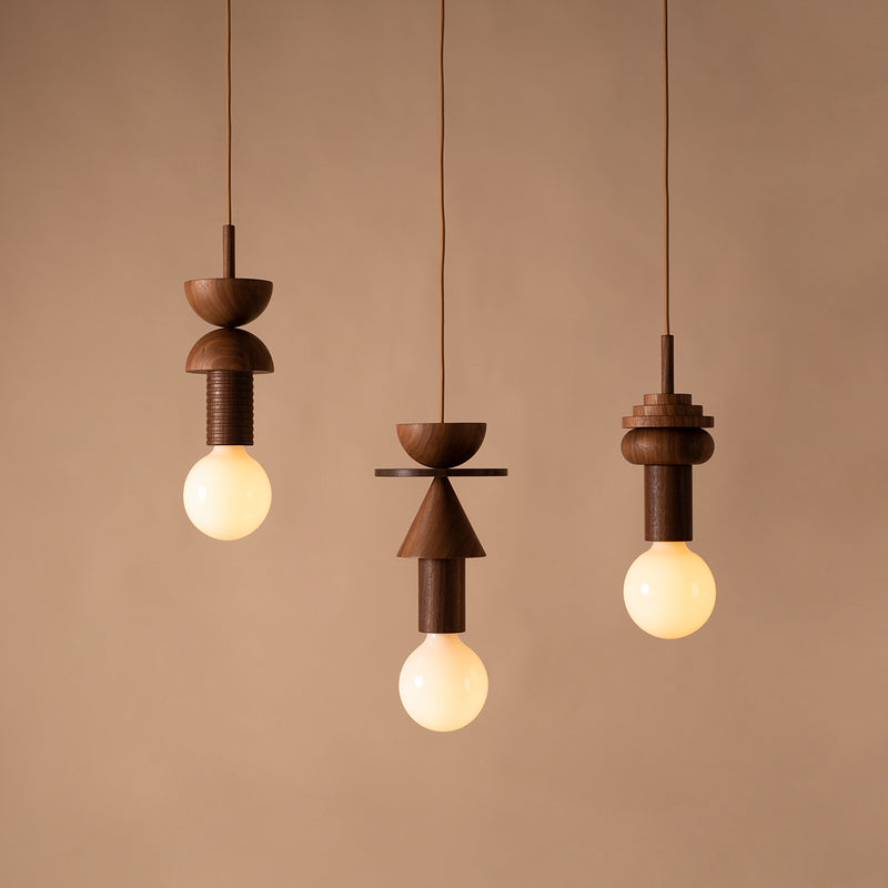 Against a beige background, three Junit Copla pendant lights by Schneid feature artistic walnut wood fixtures inspired by the Bauhaus movement. Each fixture combines spheres, cylinders, and cones in geometric harmony around a glowing bulb.