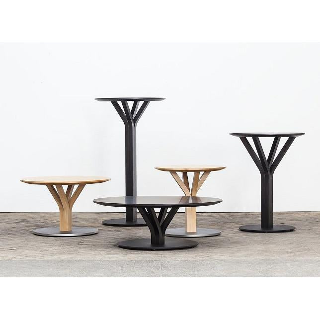 A group of Bloom Bar Table Squares by TON, featuring round tops and Y-shaped black bases, are arranged on a gray floor against a white wall. Designed by Arik Levy, these tables vary in height and size with natural oak and sleek black finishes.