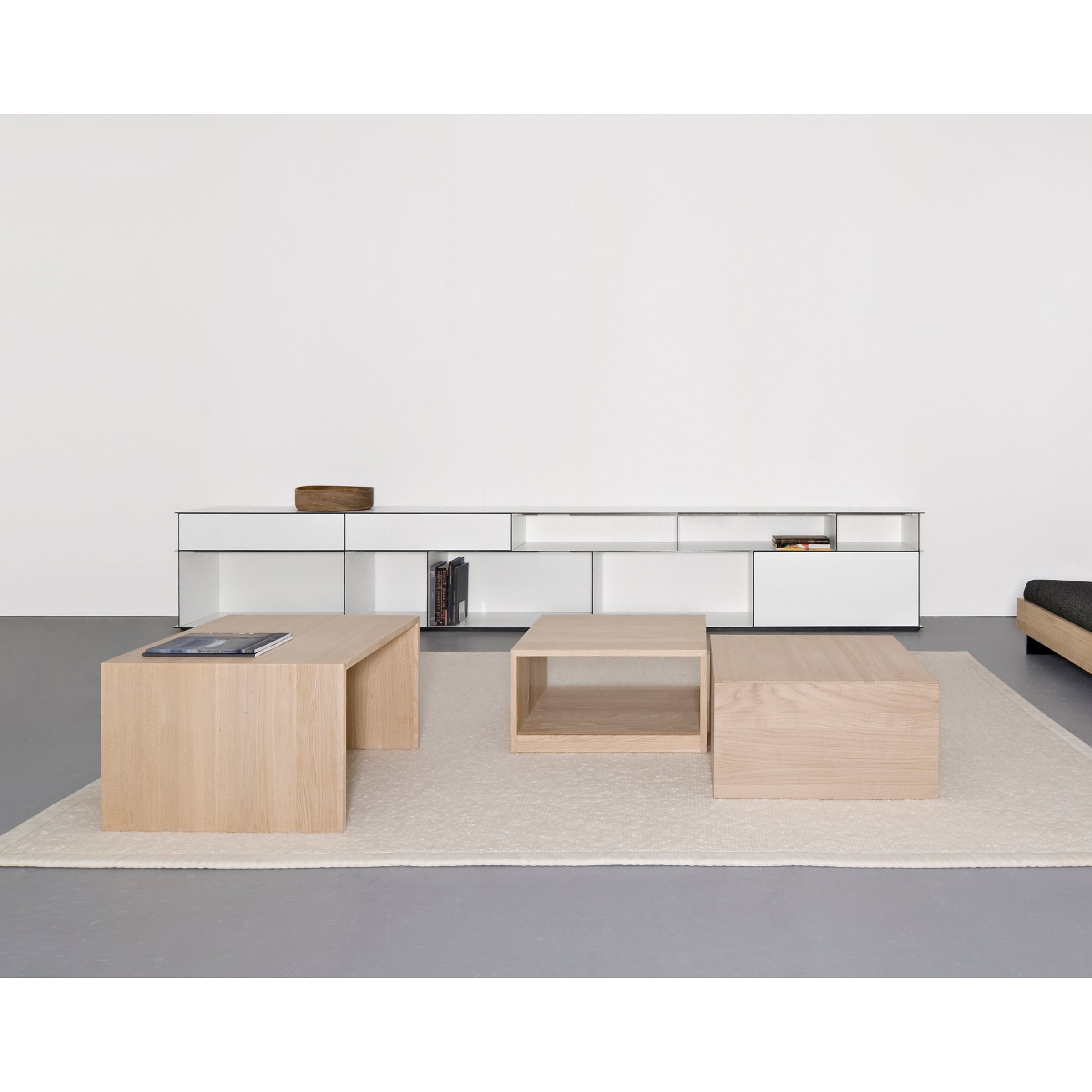 In this minimalist living room, light wood block coffee tables sit on a beige rug. A sleek low cabinet and modular sideboard from Sanktjohansers Digital multipurpose collection harmonize with the white wall, accompanied by a small wooden box and books.