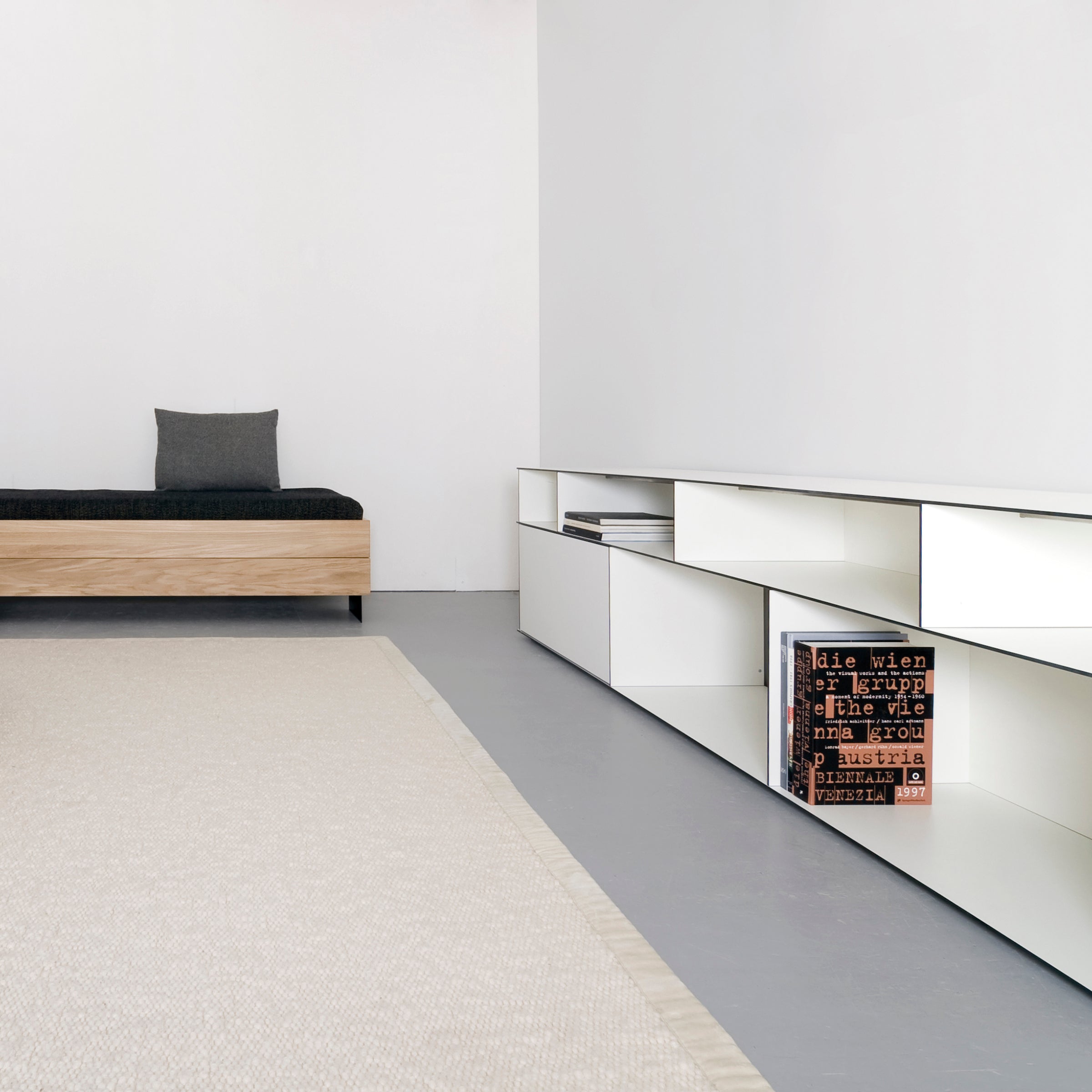 The minimalist room features a black-cushioned wooden bench against the wall. A low white shelf system showcases Sanktjohansers Digital multipurpose box adorned with text. The light gray floor pairs well with the beige textured rug extending forward.
