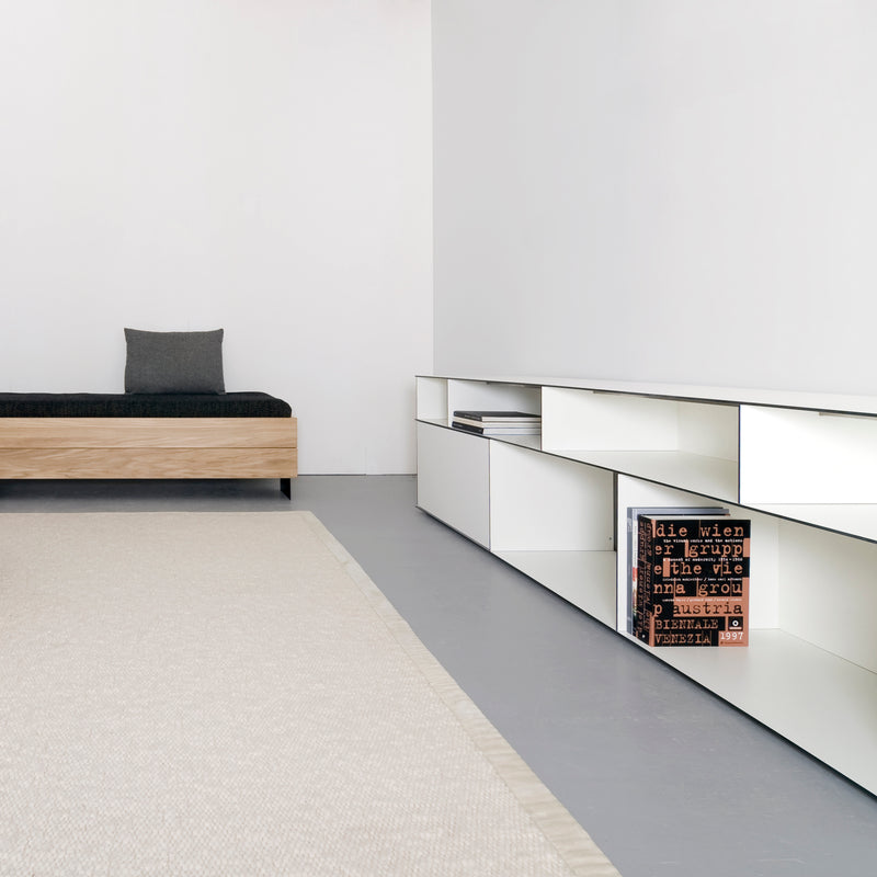 The minimalist room features a black-cushioned wooden bench against the wall. A low white shelf system showcases Sanktjohansers Digital multipurpose box adorned with text. The light gray floor pairs well with the beige textured rug extending forward.