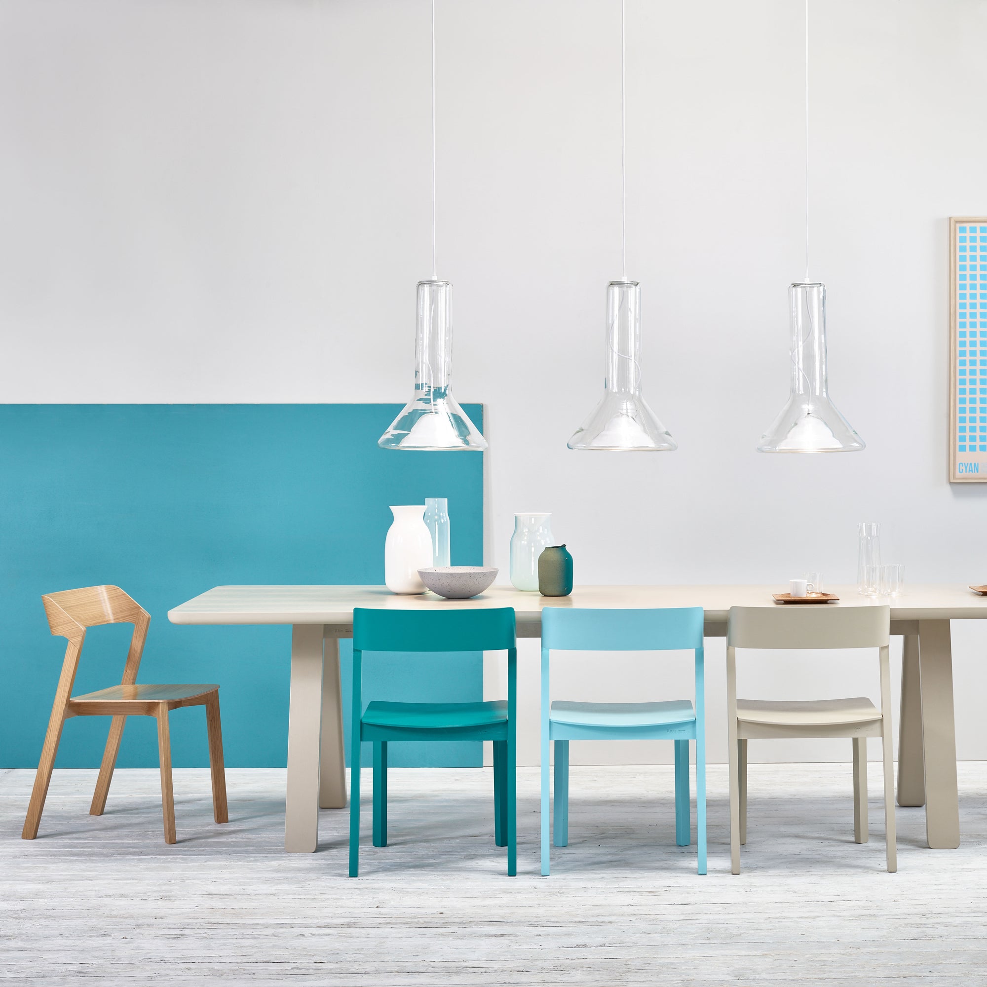 A modern dining room showcases a light wood table with Chair Merano by TON in blue, white, gray, and natural wood. The solid wood and bent plywood design adds elegance. Above, three clear pendant lights enhance minimalist decor against a white and turquoise split wall.