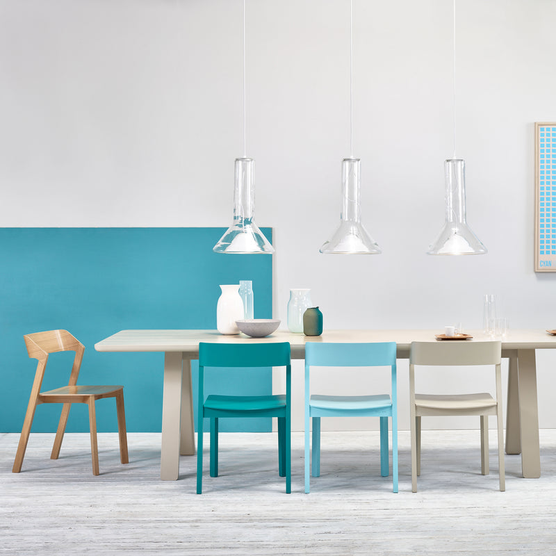 A modern dining room showcases a light wood table with Chair Merano by TON in blue, white, gray, and natural wood. The solid wood and bent plywood design adds elegance. Above, three clear pendant lights enhance minimalist decor against a white and turquoise split wall.