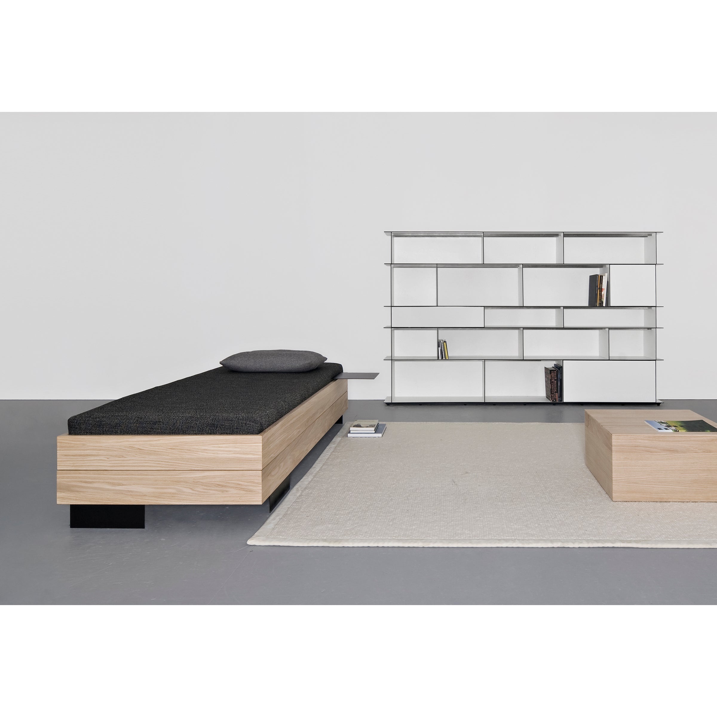 A minimalist room showcases Sanktjohansers Iku Bed Series alongside a black-cushioned wooden bench, a geometric white bookshelf with sparse books, and a solid wood coffee table on a beige rug. The gray floor and white walls enhance the serene atmosphere.