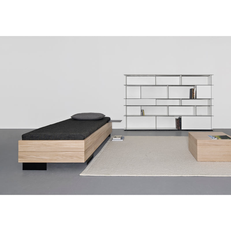 A minimalist room showcases Sanktjohansers Iku Bed Series alongside a black-cushioned wooden bench, a geometric white bookshelf with sparse books, and a solid wood coffee table on a beige rug. The gray floor and white walls enhance the serene atmosphere.