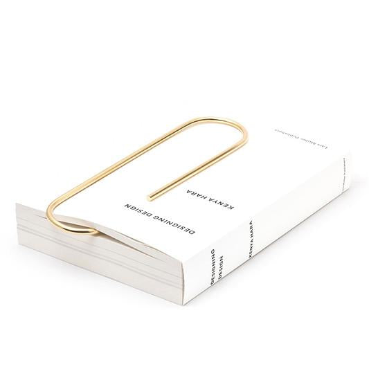 A large gold Paper Clip #4751 from Werkstätte Carl Auböck rests on a closed white book titled Designing Design by Kenya Hara. Positioned diagonally, it elegantly stretches across the book, evoking the charm of a Viennese townhouse.