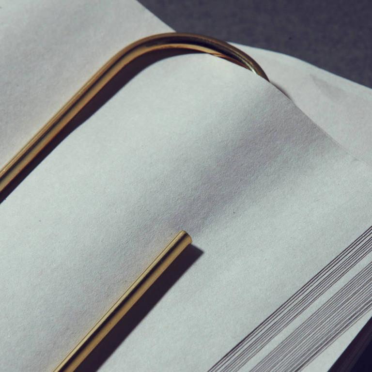 On the open book pages lies a curved, golden Werkstätte Carl Auböck Bookmark, Paper Clip #4751. Its metallic sheen catches the light, casting faint shadows on the white paper and evoking tales of elegance and timeless artistry.
