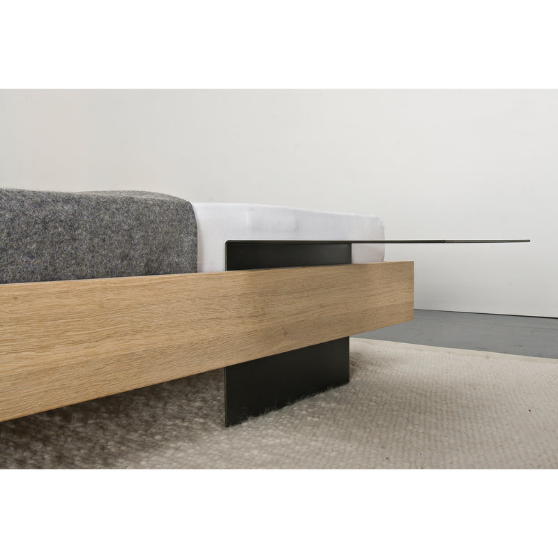 The Iku Bed Series by Sanktjohanser showcases a minimalist design with a solid wood frame and black metal legs. A dark gray blanket drapes over the white mattress, all set on a textured beige carpet against a plain white wall.
