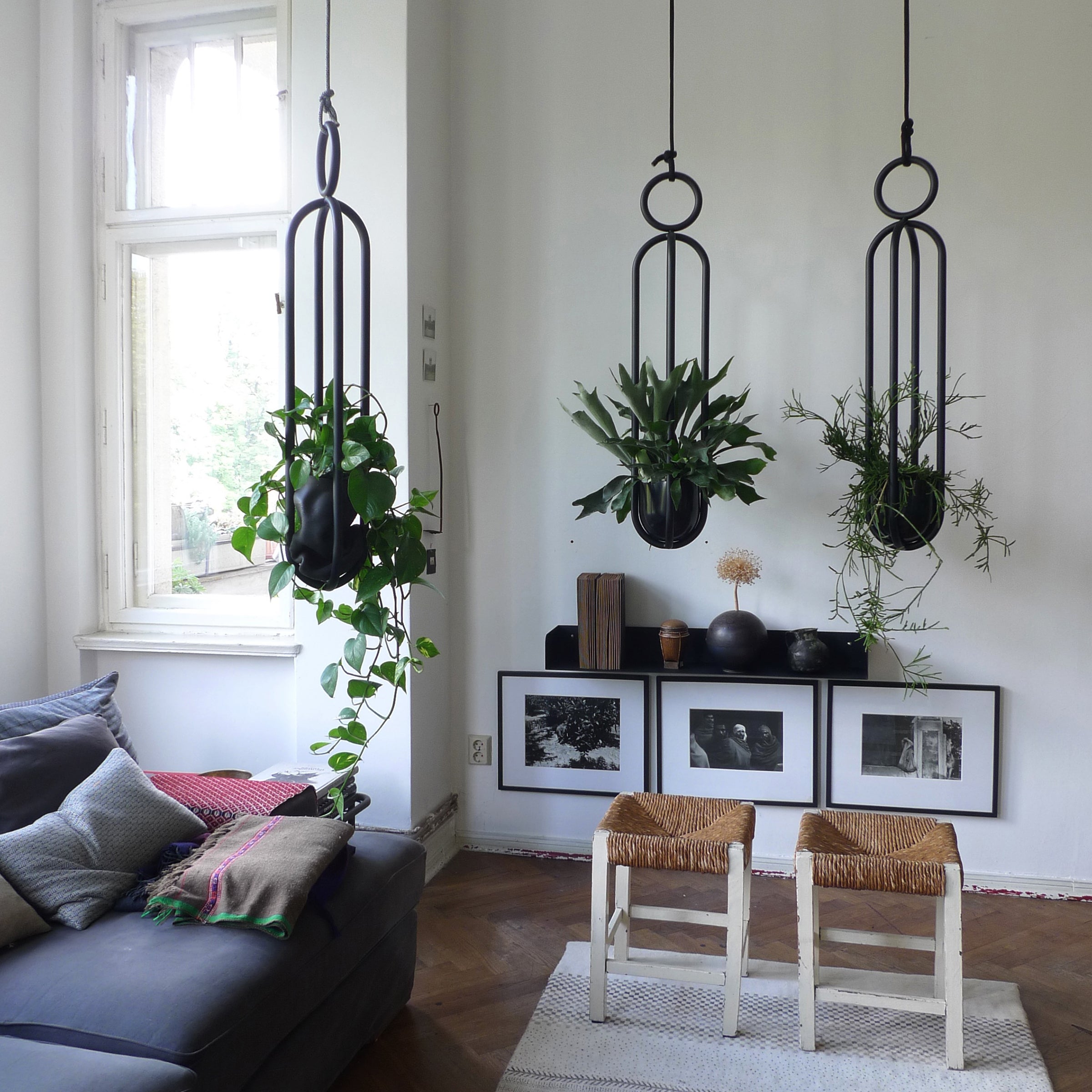A cozy living room with three Blumenampel hanging flower vases by Atelier Haussmann, a gray sofa adorned with pillows and a colorful throw, two wooden stools, and black-and-white framed photos on the wall. Natural light streams in through a large window.
