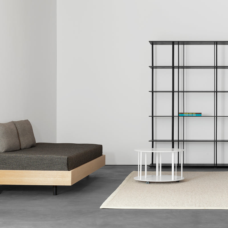 A minimalist living room features a Sanktjohanser Iku Bed Series daybed with dark cushions, resembling sleek bed frames, complemented by a thin black bookshelf with sparse books. A round white side table rests on a light rug, all unified by the room’s neutral palette and clean lines.
