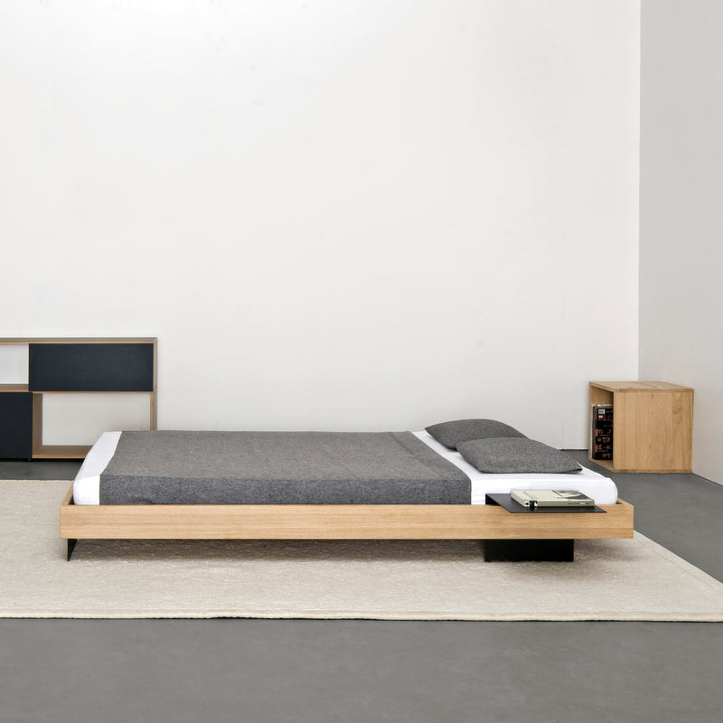 A minimalist bedroom includes the Iku Bed Series by Sanktjohanser, featuring a low solid wood frame with gray bedding and a built-in side table. A light carpet covers the gray floor, while a wooden and black shelving unit stands against a pristine white wall in the background.