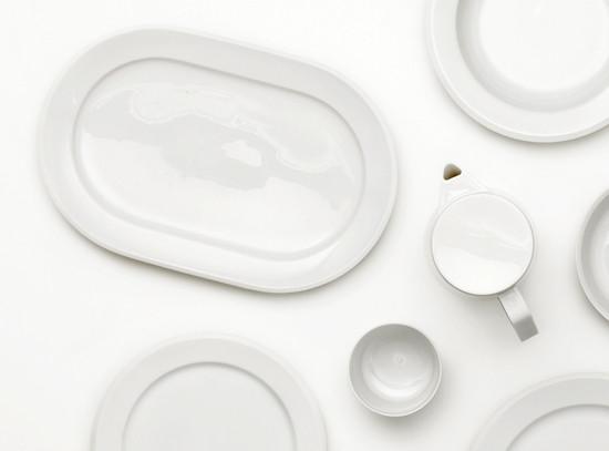 The Augarten Orbit Collection features a minimalist arrangement of white porcelain tableware on a white surface, including an oval platter, handled pitcher, plates, and bowls. Highlighted by its rotational shapes and semi-matt polished rims, the collection is currently on sale.