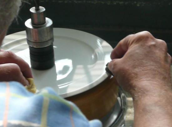 In a blue plaid shirt, a person skillfully engraves elegant shapes onto the edge of a white plate from the Augarten Orbit Collection - Sale. Sunlight filters through the window, illuminating their work on the semi-matt polished rim of this exquisite porcelain by Augarten.