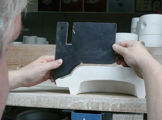 A person uses a black L-shaped tool for measuring a white sculpted object, likely part of crafting the Augarten Orbit Collections exquisite porcelain tableware. Several semi-matt polished white cylindrical pieces are on a wooden shelf in the background, all illuminated well.