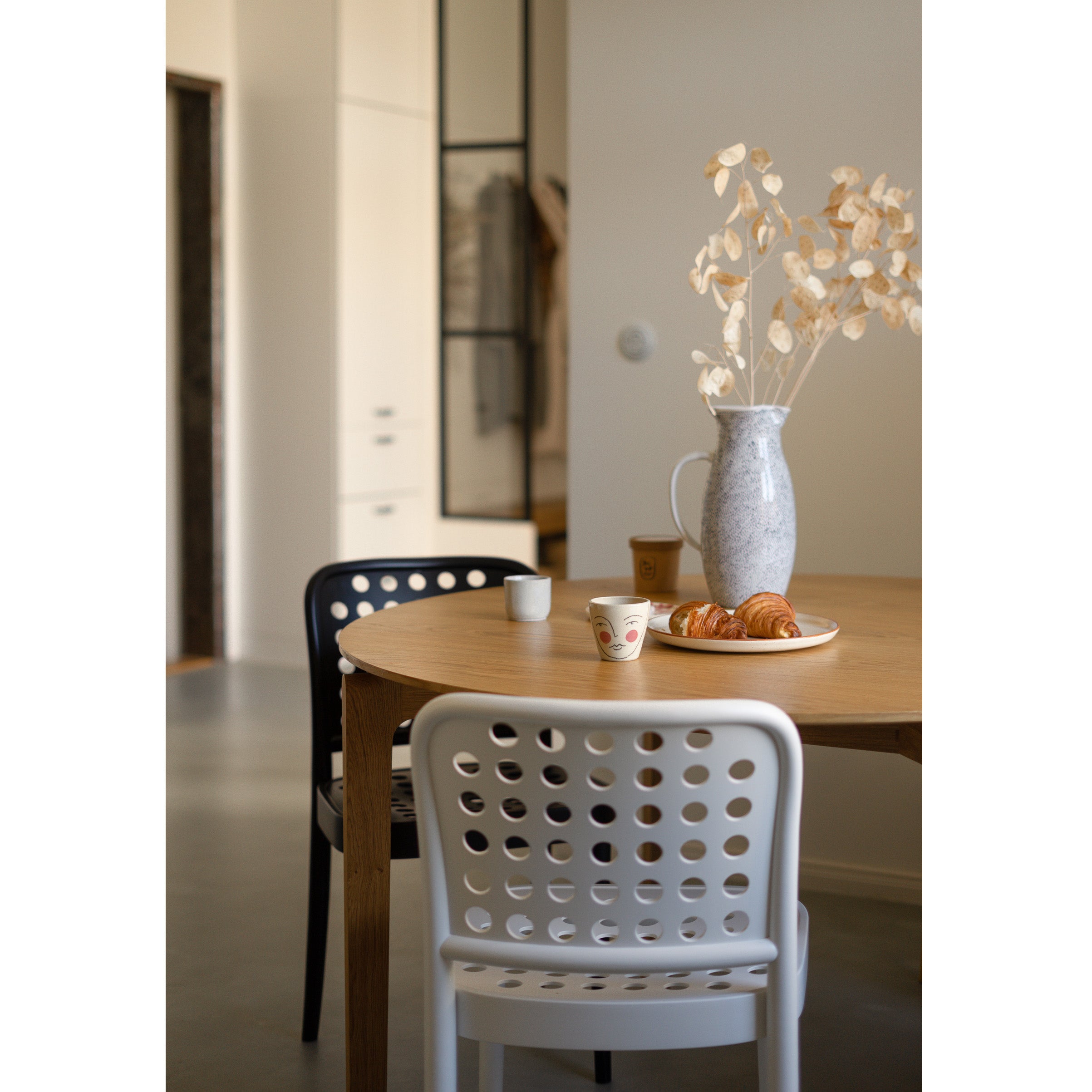 A cozy dining area perfectly captures Scandinavian minimalism with a wooden table and TONs Chair 822 in black and white. A grey vase with dried flowers, croissants on a plate, and cups sit on the table. The background features a sleek, minimalistic kitchen.