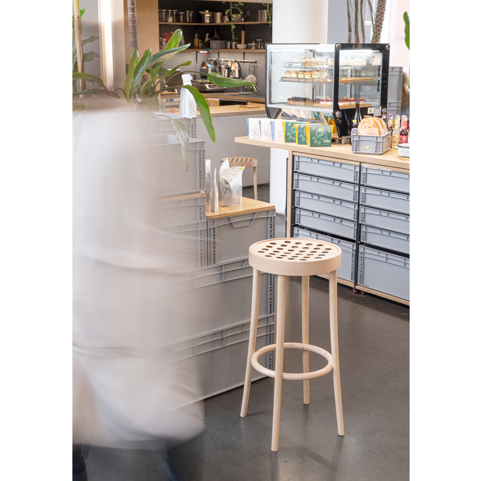 The Barstool 822 by TON, made with manual wood-bending technology and a perforated seat, sits beside gray boxes in a café. A persons motion creates a blur effect, while the counter showcases pastries in a glass case and tea boxes.