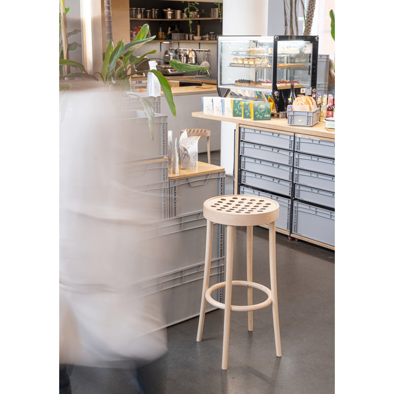 The Barstool 822 by TON, made with manual wood-bending technology and a perforated seat, sits beside gray boxes in a café. A persons motion creates a blur effect, while the counter showcases pastries in a glass case and tea boxes.