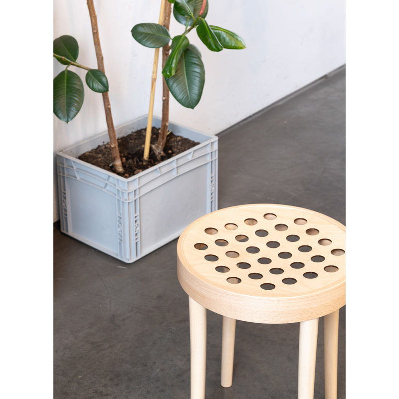 The Stool 822 by TON, featuring a perforated seat, highlights Scandinavian minimalism on a gray floor. Beside it sits a plant with broad leaves in a gray crate-like pot designed by Claesson Koivisto Rune, all beautifully arranged against a white wall.