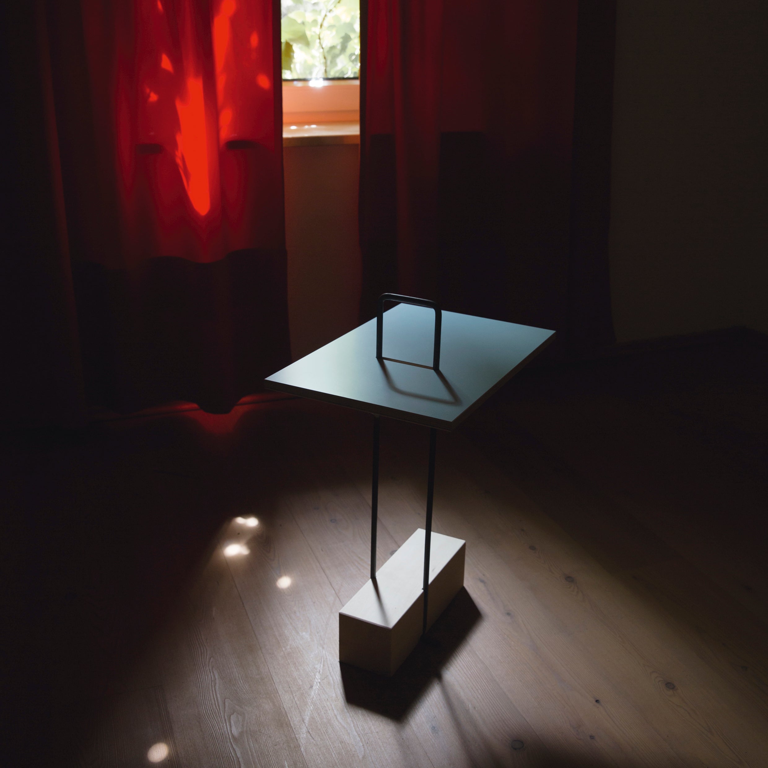 The Abgemahnt side table by Moormann, designed by Nils Holger Moormann, has a minimalist look with a thin metal frame and a small rectangular top. It is set on a wooden floor in a dimly lit room with red curtains drawn, letting the diverse tabletop hues catch the filtered natural light.