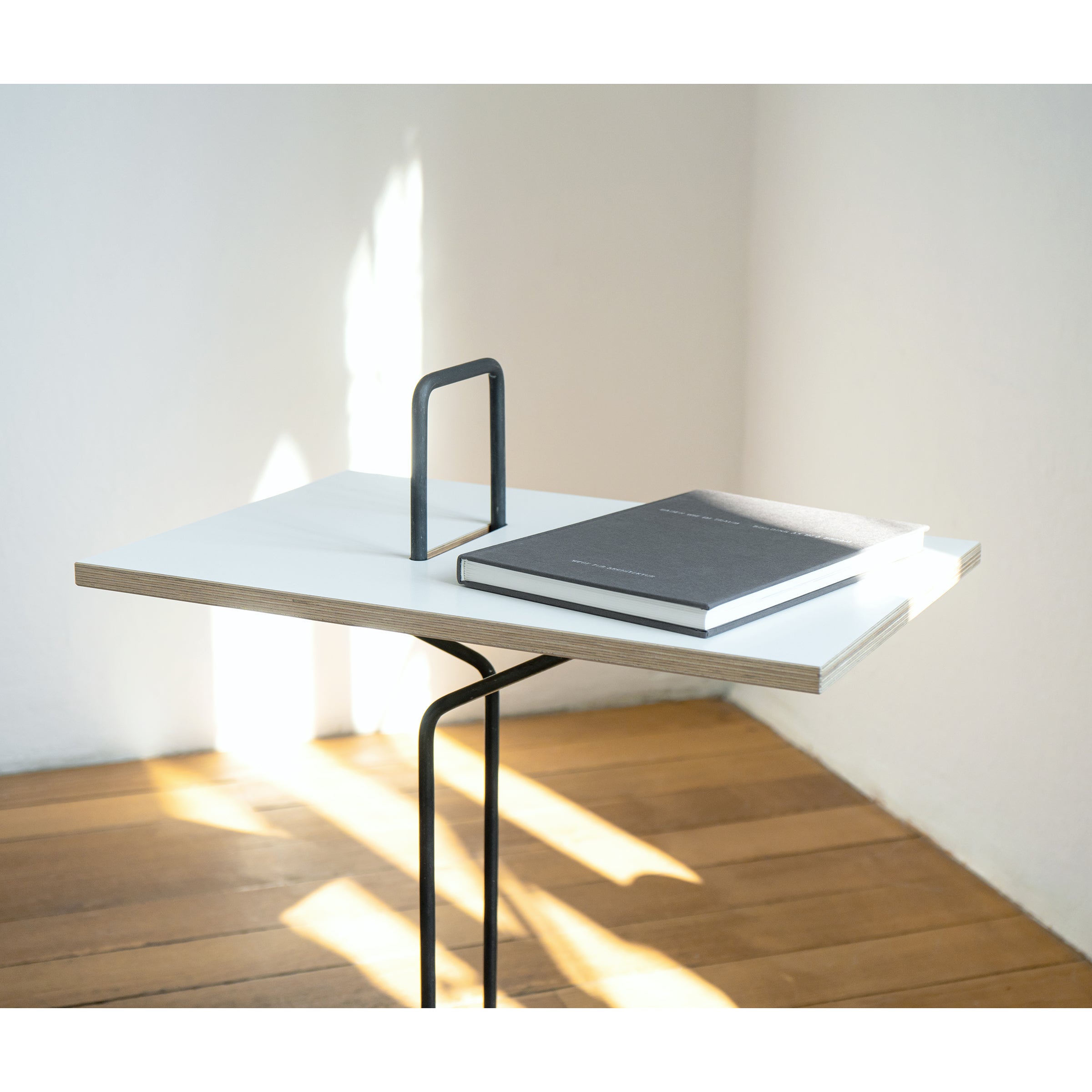 A minimalist Moormann Abgemahnt side table with a white top and slim metal legs stands by a white wall. A closed black notebook sits on the table, while sunlight dances across the wooden floor, casting soft shadows.