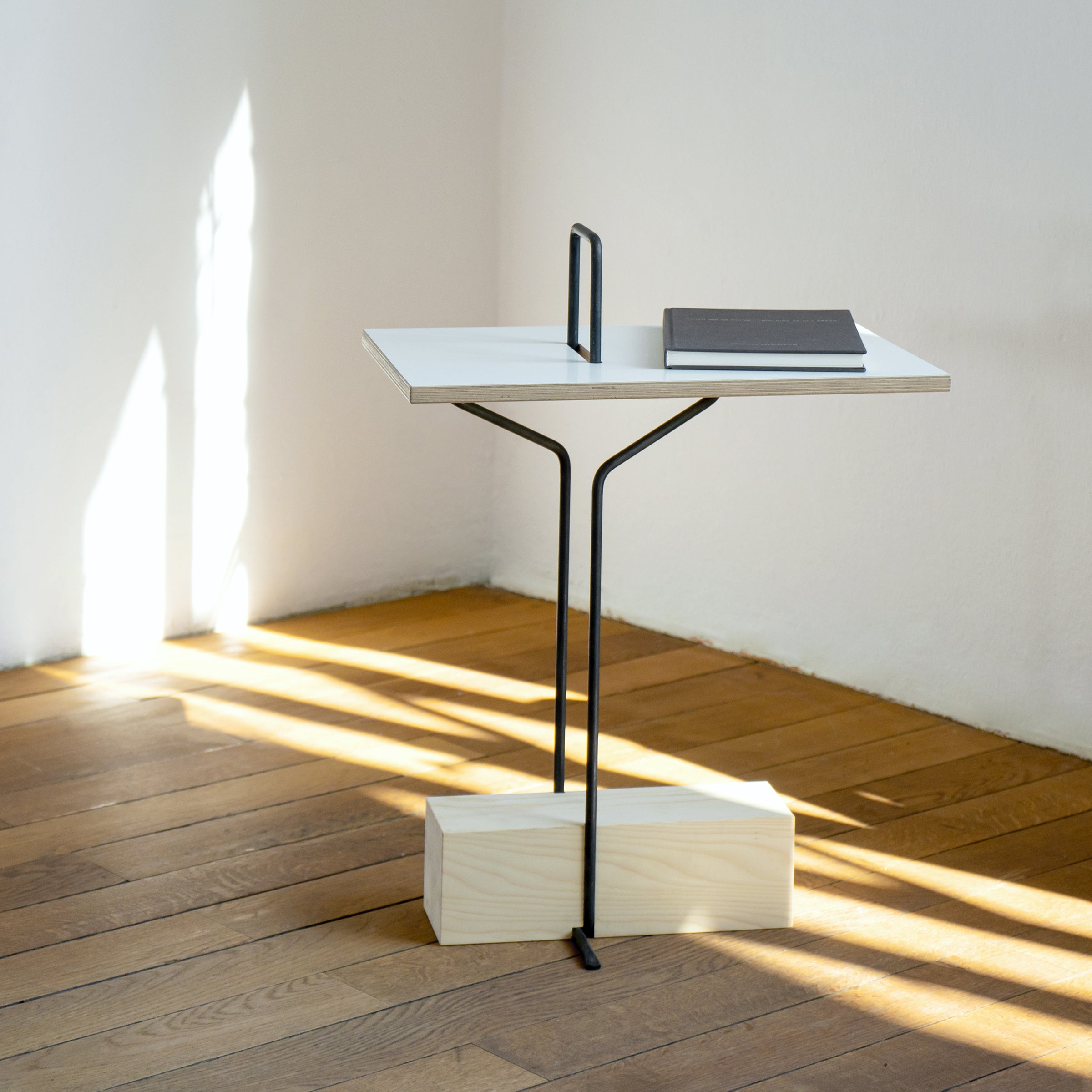 The Abgemahnt side table by Moormann showcases a minimalist design with a thin black metal frame and white rectangular top, enhancing the tabletops colors. Sunlight creates shadows on the wooden floor as a closed, dark-colored book sits quietly atop.