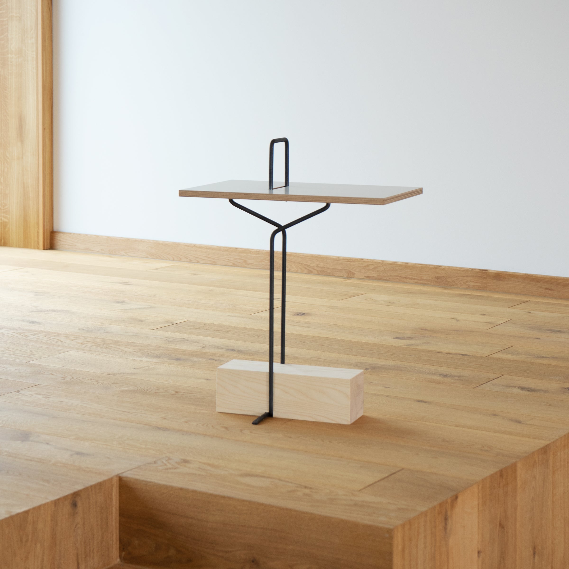 The Abgemahnt by Moormann is a modern minimalist side table with a wooden top in various colors and a sleek black metal base anchored to a rectangular wooden block. It sits elegantly on a light wood floor against a white wall.