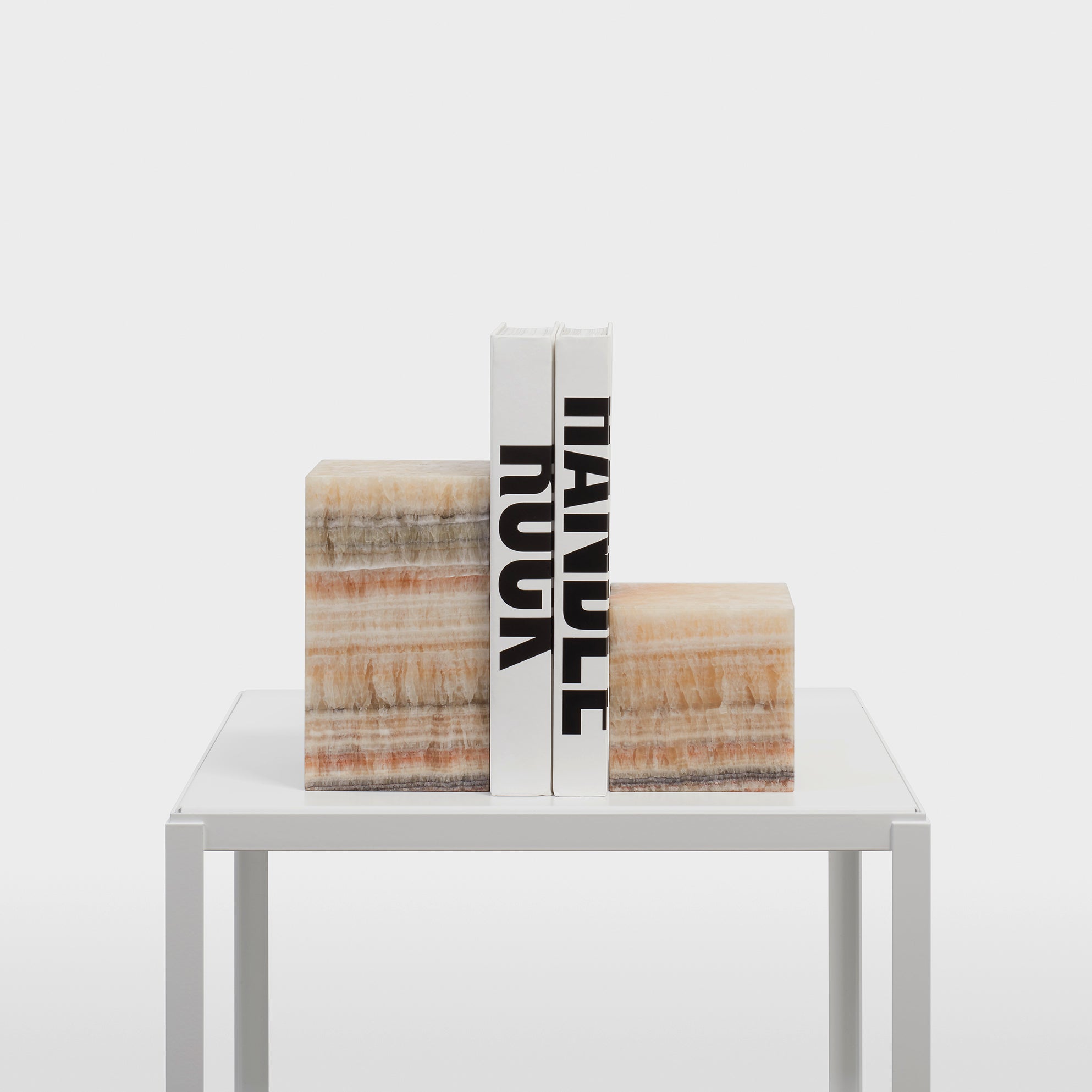 Two books titled HANDLE and ROCK stand vertically on a white table, supported by two AC11 STOP ONYX bookends with natural brown and cream striations. These sculptural accessories from the E15 collection add elegance against the plain white background.