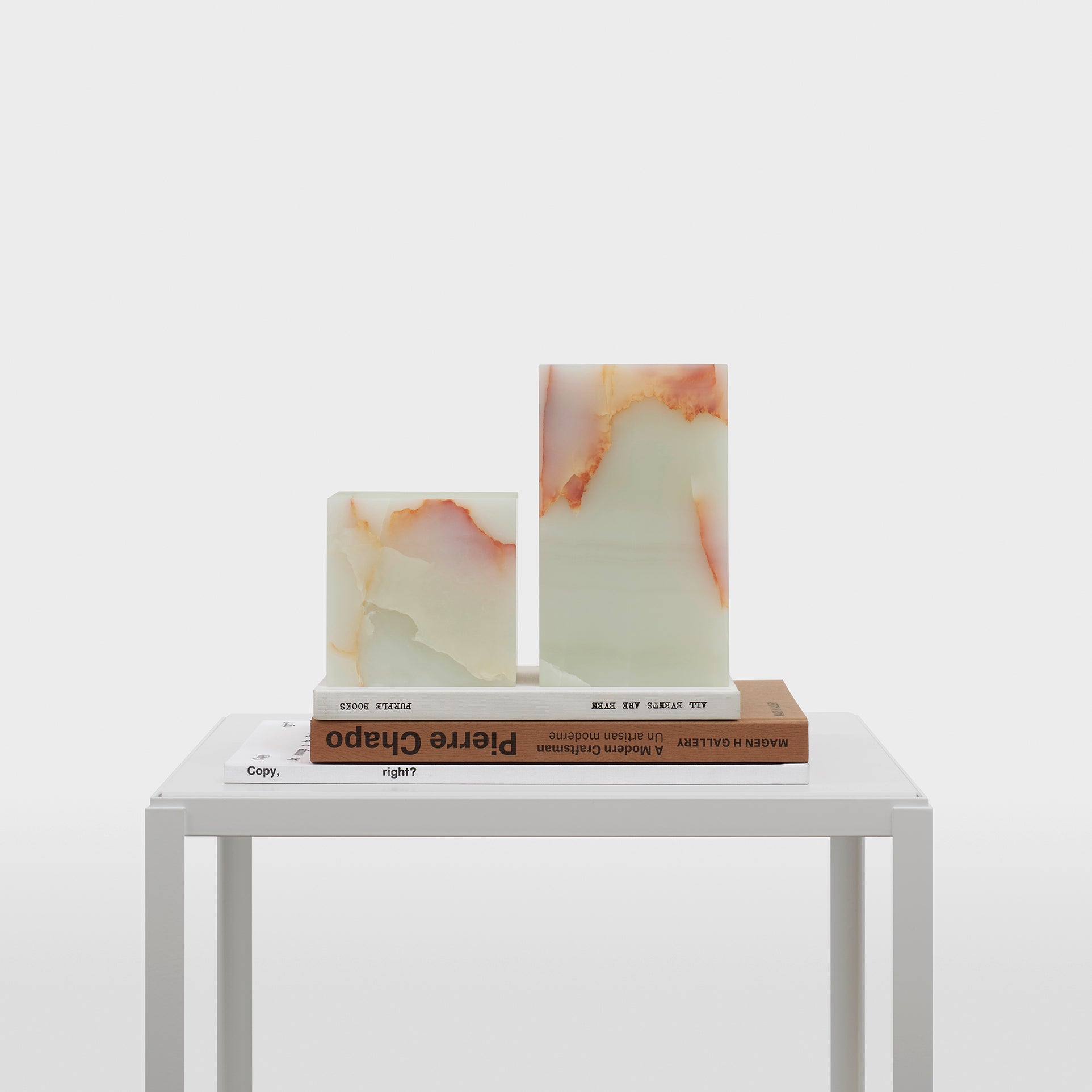 The AC11 STOP ONYX marble bookends by E15, in elegant earth tones, hold stacked books on a simple white table. The minimalist white background enhances the modern aesthetic of this sculptural accessory.