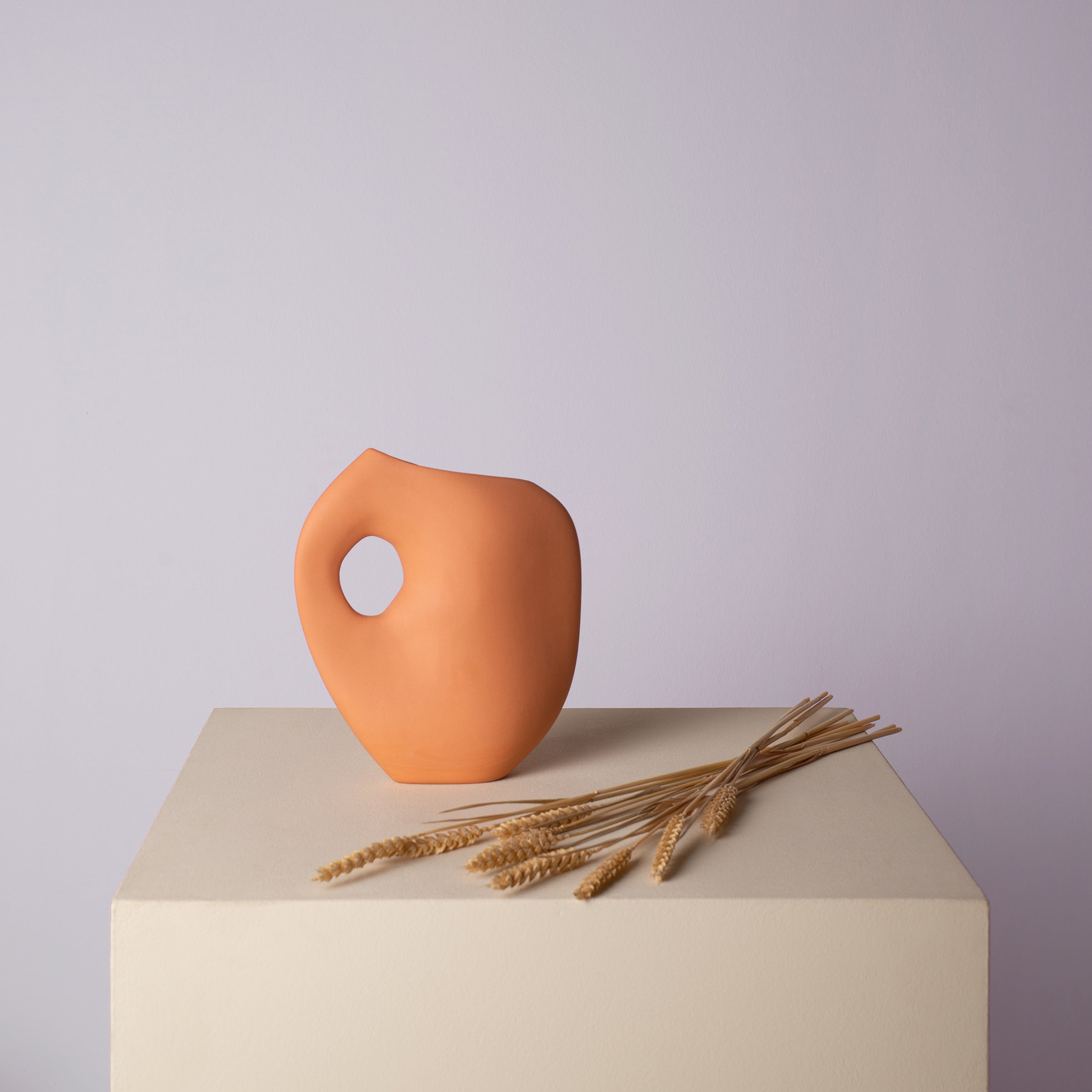 An orange ceramic vase with a looped handle from Schneids Aura I collection stands on a beige pedestal, accompanied by dried wheat stalks and set against a muted lavender background.