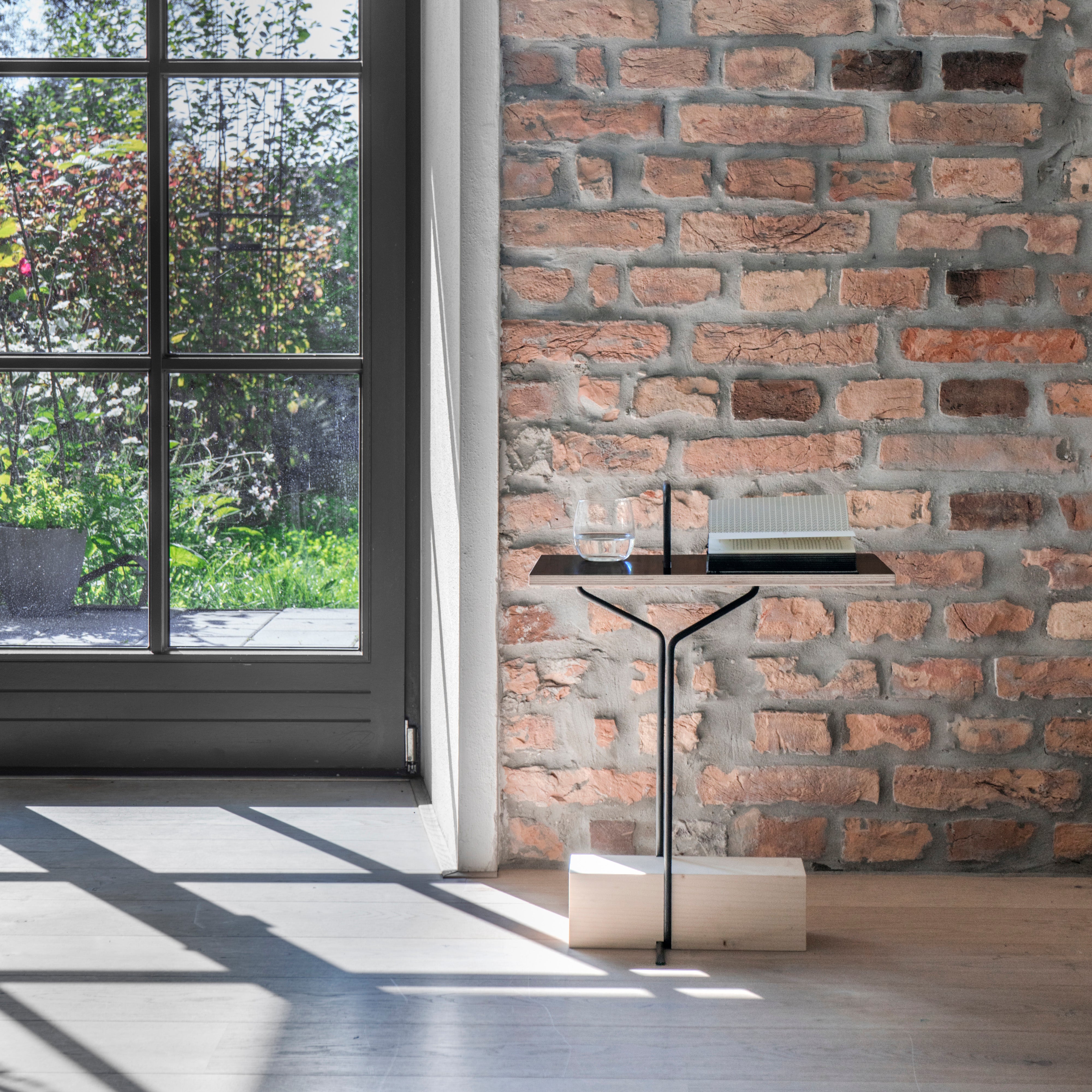 In a sunlit room with brick walls, Moormanns Abgemahnt side table features a colorful glass top on a wooden block, holding an open book and a glass of water. A large window offers views of greenery outside, casting shadows on the tabletop and wooden floor.