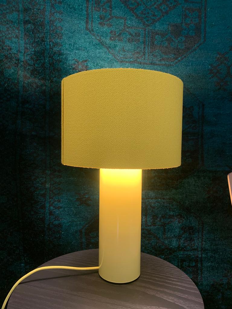 The ALL ROUND MINI Table Lamp - SALE by Victor Foxtrot is a stylish lamp with a cylindrical yellow wool fabric shade, producing a soft, warm glow on a dark wooden surface against an elegantly patterned teal backdrop.