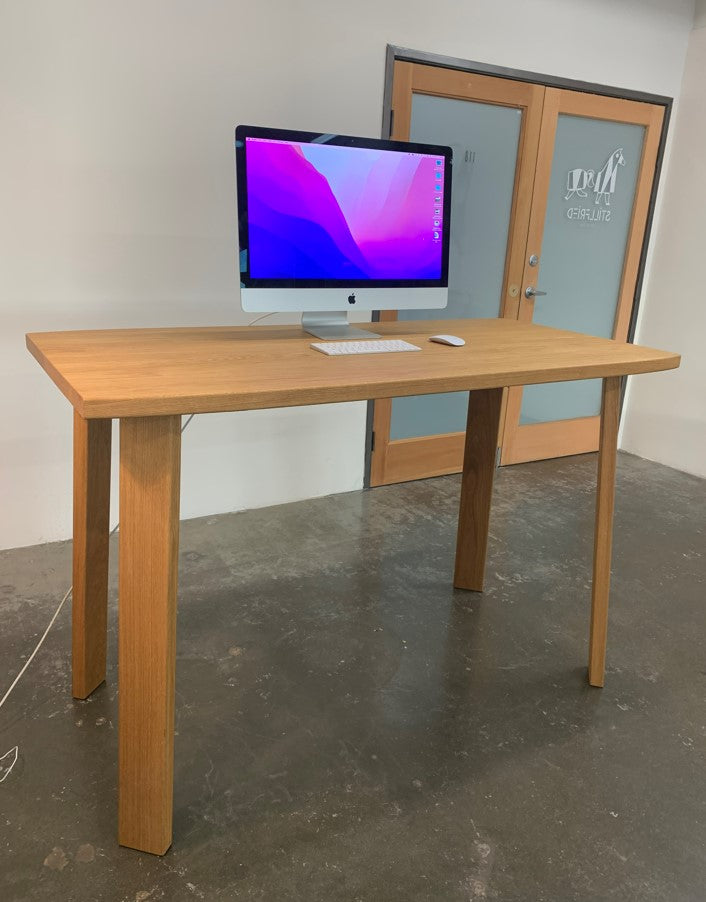 The Alpin Bar Table by Hussl from the Alpine collection showcases a wooden surface with a computer monitor, keyboard, and mouse on it. Positioned in an office with a frosted glass door, it reflects the refined simplicity of EOOS设计.