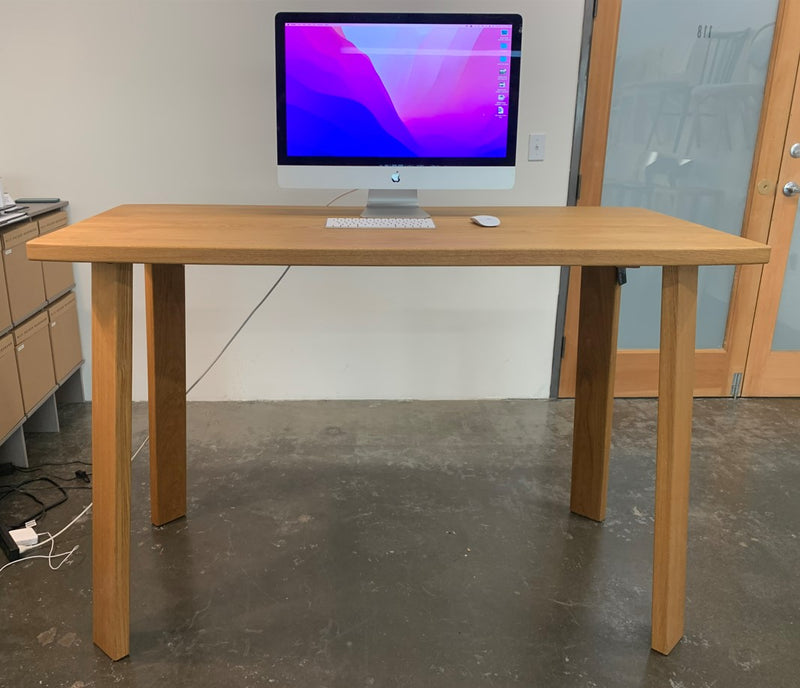 The Alpin Bar Table - Sale by Hussl showcases a sleek design, complete with a modern desktop computer and mouse. This Alpine piece rests on polished concrete, with light streaming through a nearby frosted glass door.