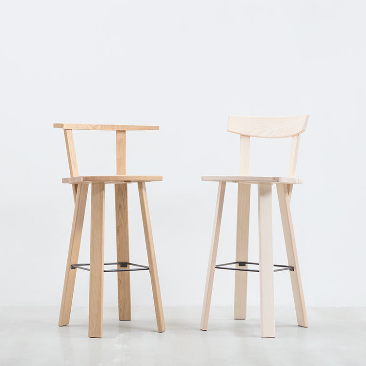 Two Alpin Barstool BSO from Hussl with minimalist designs, a low backrest, and black metal footrests stand against a gray backdrop. The left stool features darker wood, while the right showcases a lighter shade.