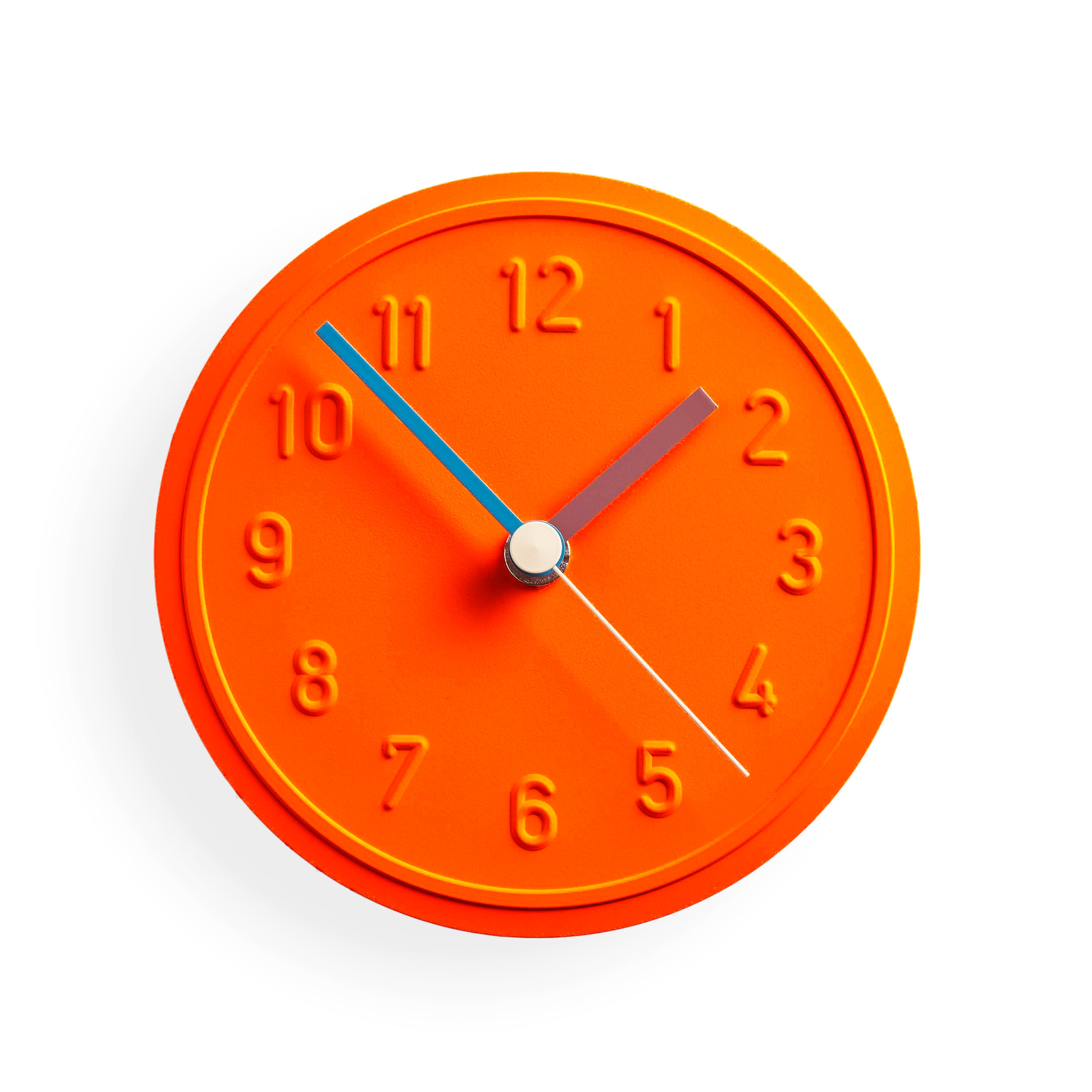 The Alu Alu Wallclock by Richard Lampert, designed by Jochen Gros, is an orange clock with embossed numbers. It has red, blue, and white hands for hours, minutes, and seconds respectively. This elegant quartz clock shows 2:10 on a plain white background.